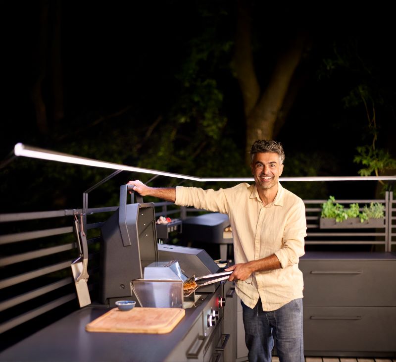 Man smiling while grilling food on an outdoor barbecue grill at night on a patio.