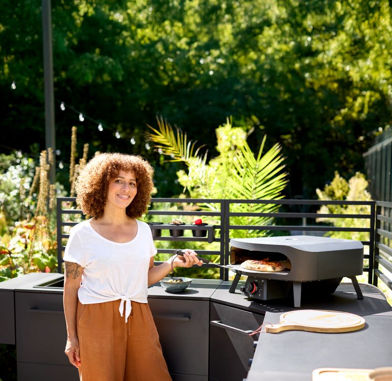 Woman using a pizza oven on an outdoor kitchen counter, pulling out a pizza on a peel, surrounded by greenery.