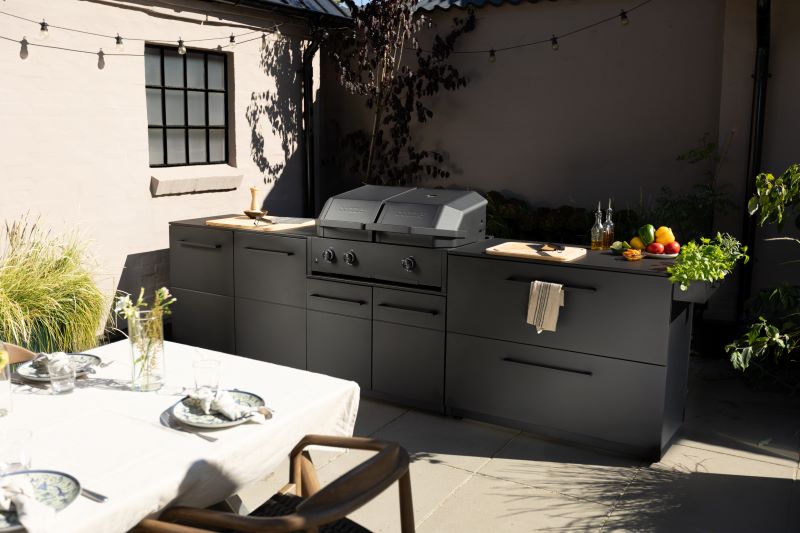 Modern outdoor kitchen setup with a black grill, countertop, cutting boards, utensils, and fresh vegetables in a sunlit patio area.