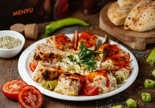 Plate of Turkish-style baked stuffed flatbread rolls, garnished with fresh parsley and dried herbs, surrounded by sliced tomatoes and green chili peppers, with more flatbreads on a wooden board in the background.