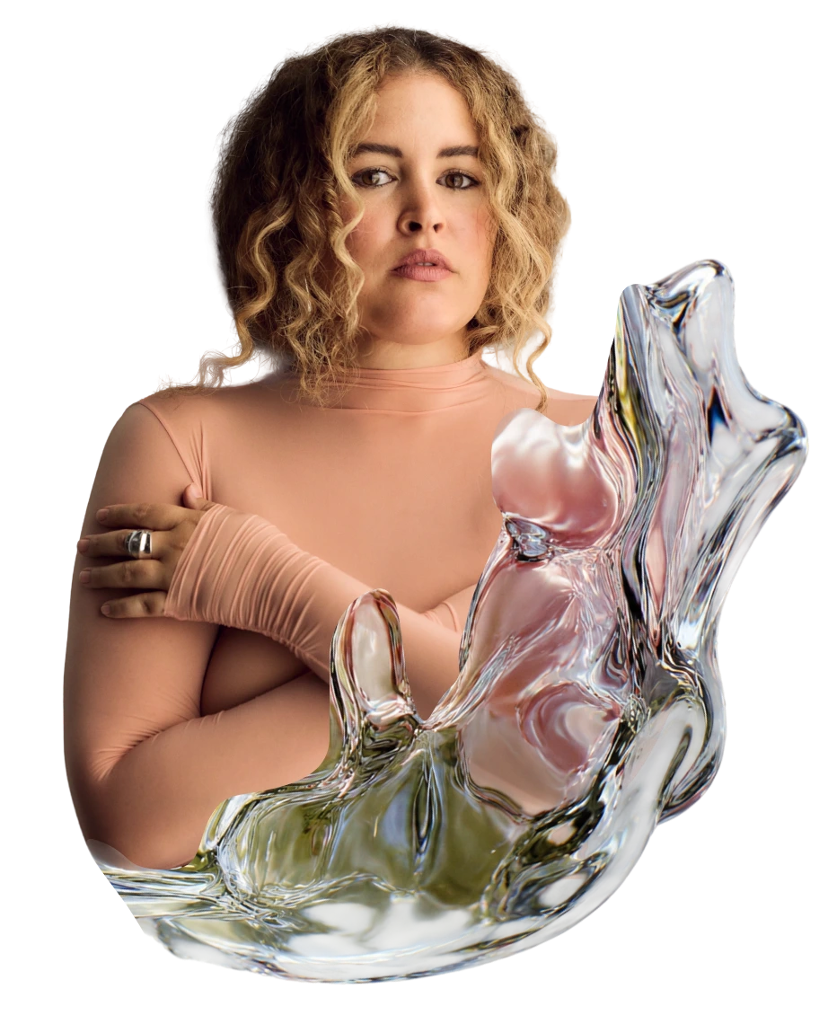Susana with curly hair wearing a sheer peach outfit and gloves, seated behind a large reflective glass sculpture against a light background.