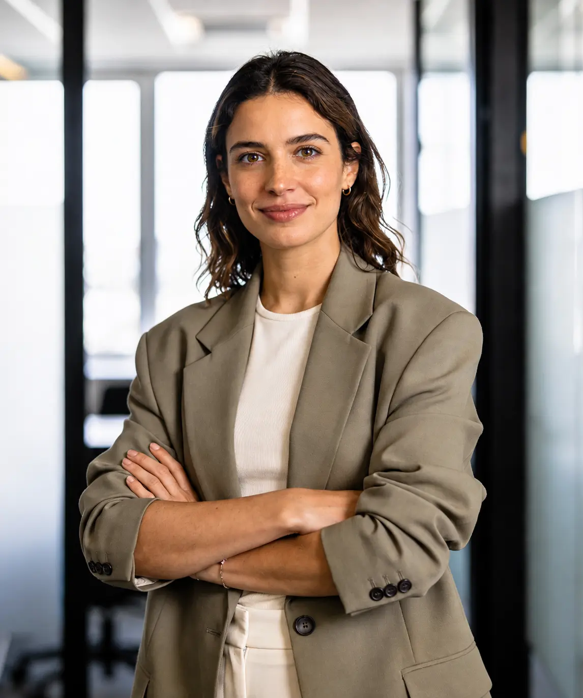 Confident woman in a beige blazer with arms crossed, standing in a modern office.