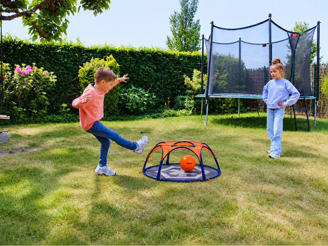 A boy kicks an orange ball at the Pannaball goal in a garden while a girl watches, with a trampoline in the background