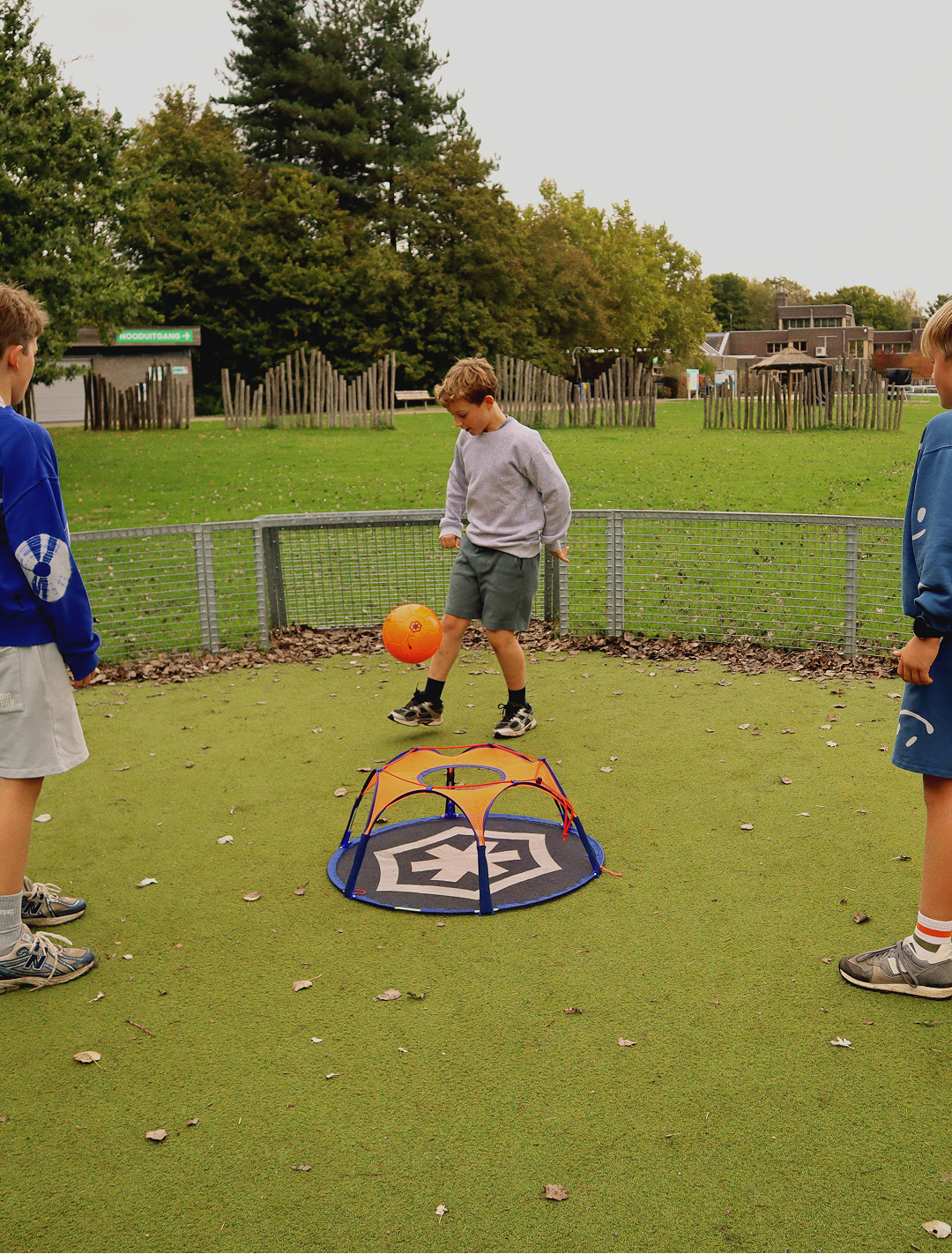 A boy juggles an orange ball above the Pannaball goal in a fenced panna court on artificial turf, two friends watching