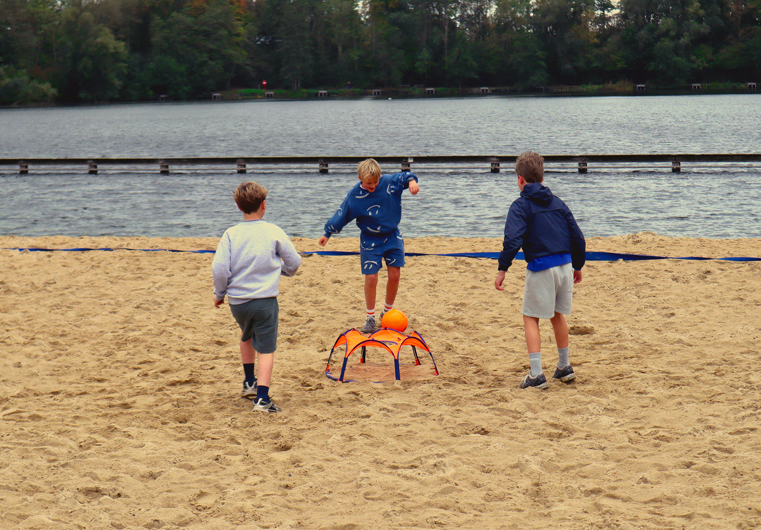 Three boys playing panna on the sand by a lake, with the orange Pannaball goal and ball in the middle