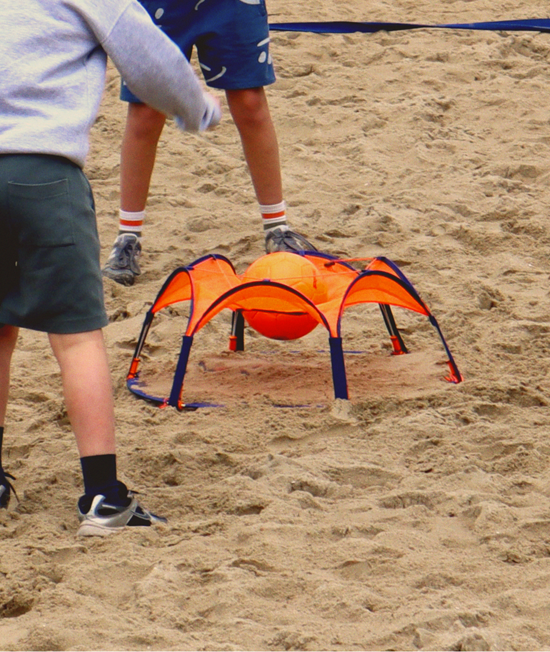 Close-up of the Pannaball goal in the sand with an orange ball inside, two boys ready around it