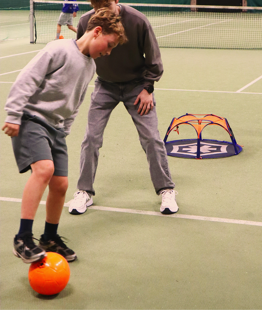 A boy dribbles an orange ball past an older player in a sports hall, with the Pannaball goal in the background