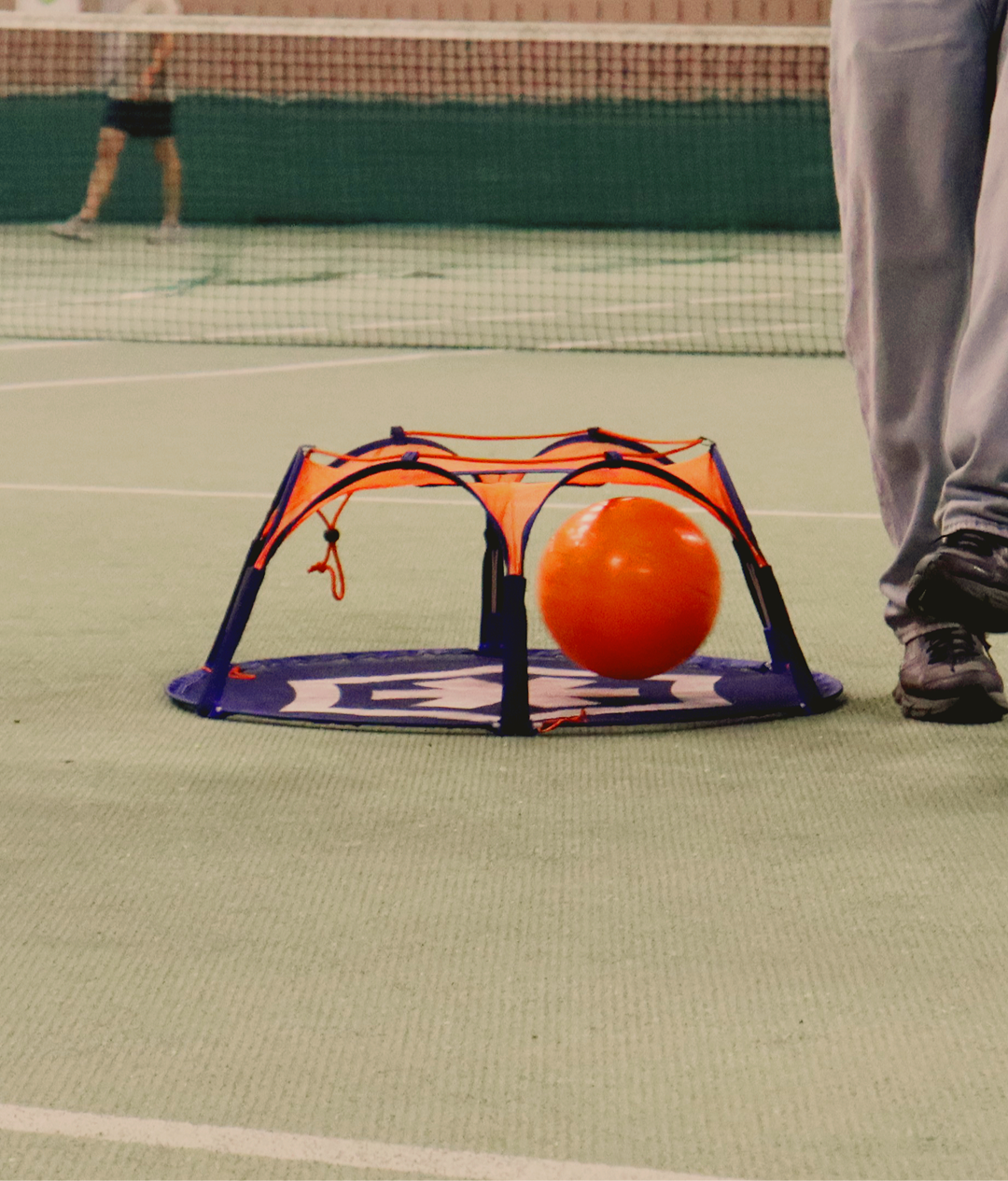 Close-up of the Pannaball goal with an orange ball inside on a green indoor sports floor, a player stepping past