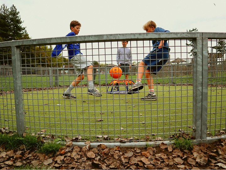 Three boys playing soccer on a small fenced grassy pitch; two are kicking an orange soccer ball towards a Pannaball goal while the third watches inside the enclosure.
