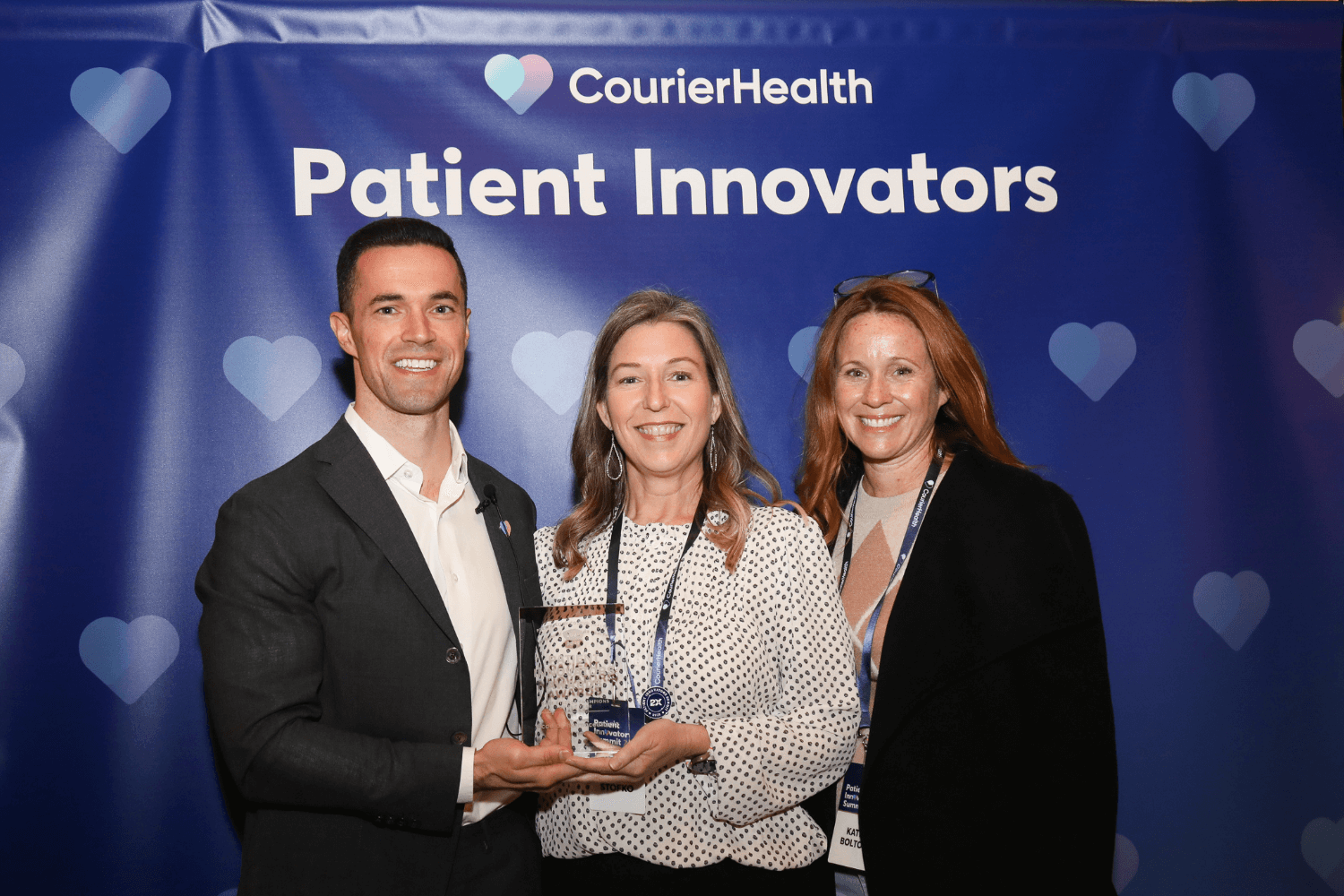 Three people smiling in front of a CourierHealth Patient Innovators backdrop; the man on the left in a dark suit and white shirt and the woman in the middle hold a transparent award together, while the woman on the right, wearing a black jacket, stands next to them.