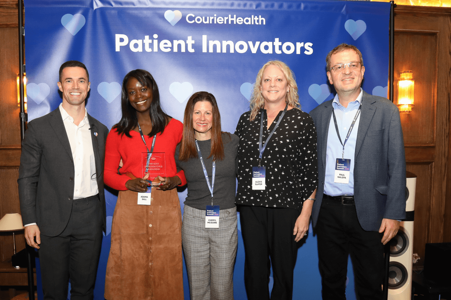 Five people standing in front of a CourierHealth Patient Innovators backdrop, smiling at the camera. The second person from the left, wearing a red top and brown skirt, holds a clear award plaque. All are wearing event badges with names visible: Adama Sali, Cheryl McGuire, Alexis Slater, and Paul Nalepa.