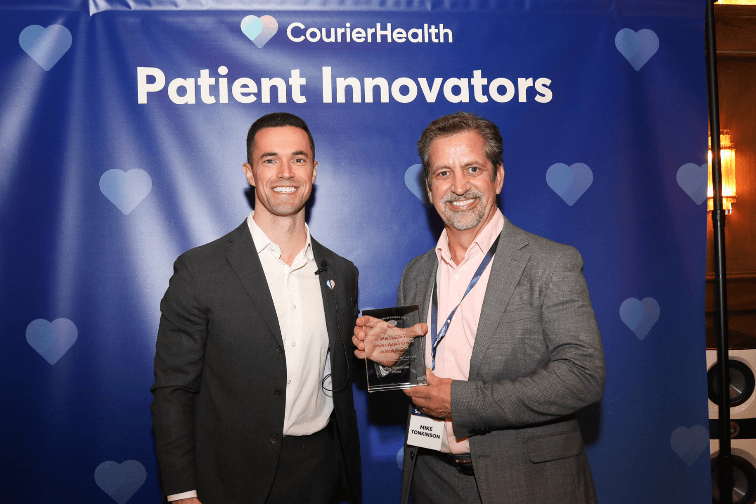Two men in business attire stand in front of a blue CourierHealth banner that reads 'Patient Innovators'; the man on the right holds a transparent award with both hands and wears a name badge reading 'Mike Tonkinson'.