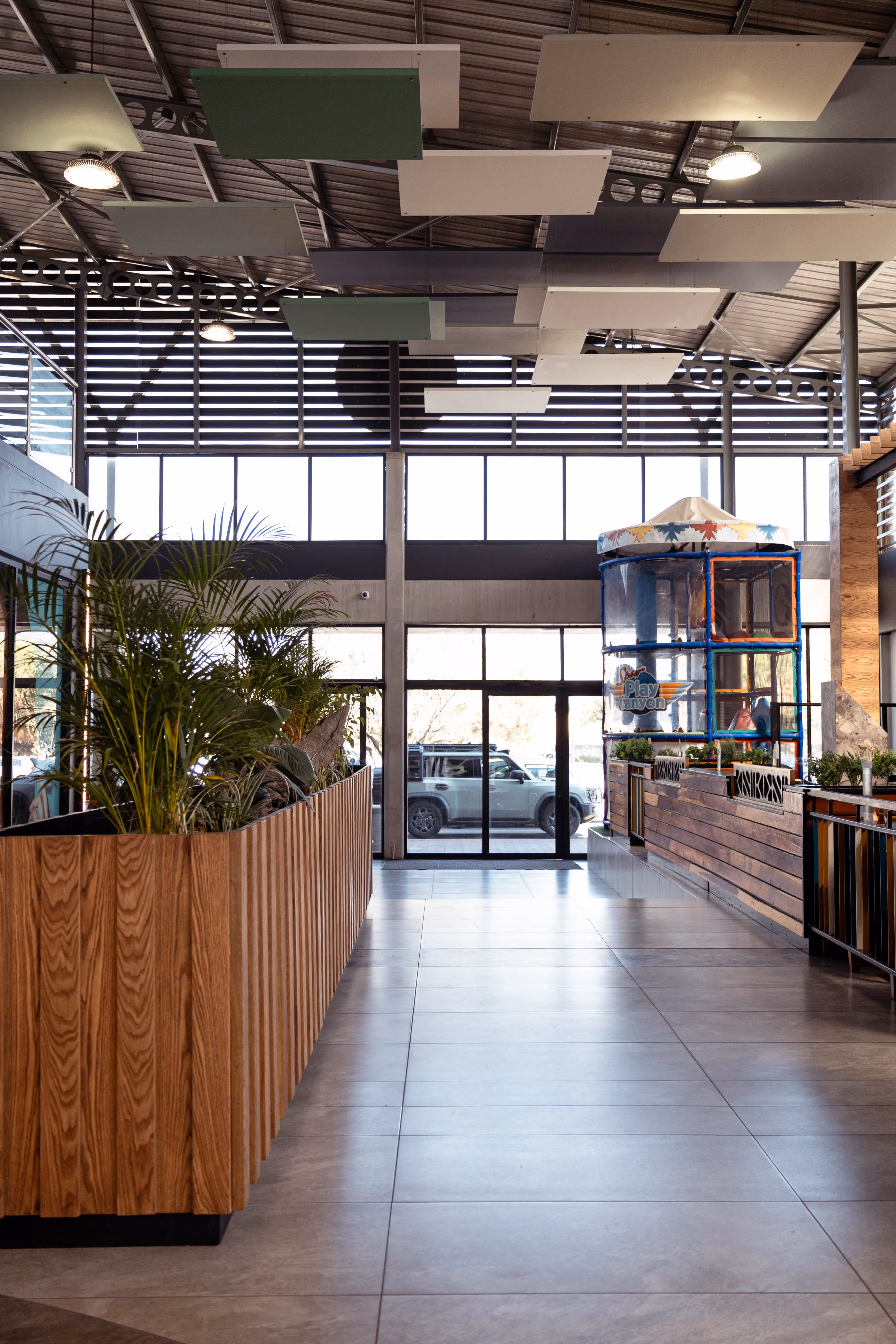 Indoor space with large windows, plants in a wooden planter, and a colorful play area near the entrance.