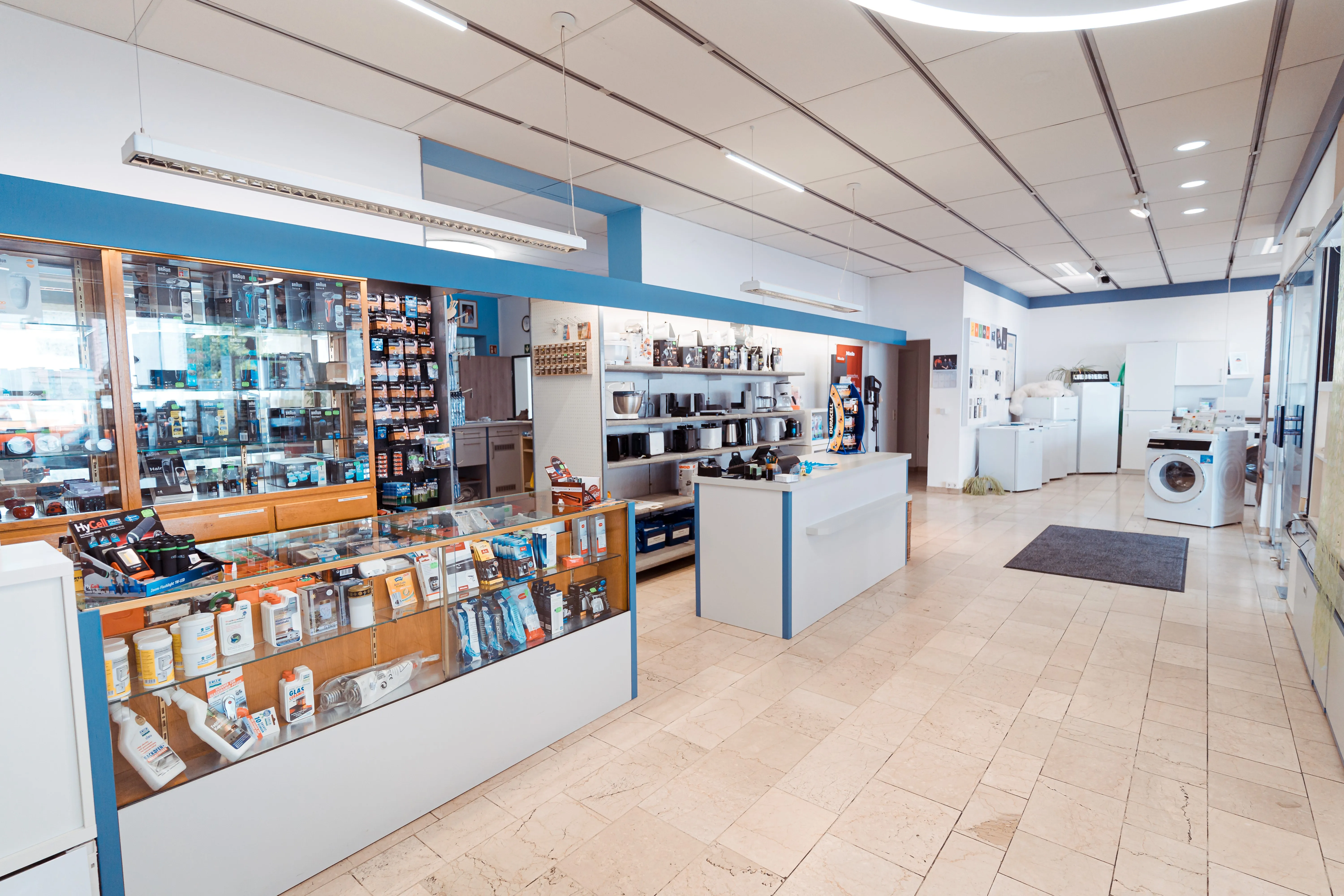 Innenaufnahme eines gut beleuchteten Elektroladens mit weißen Fliesenboden, einem Glastresen mit verschiedenen Elektronikartikeln und Regalen im Hintergrund mit Haushaltsgeräten wie Kaffeemaschinen, Kühlschränken und einer Waschmaschine rechts im Raum.