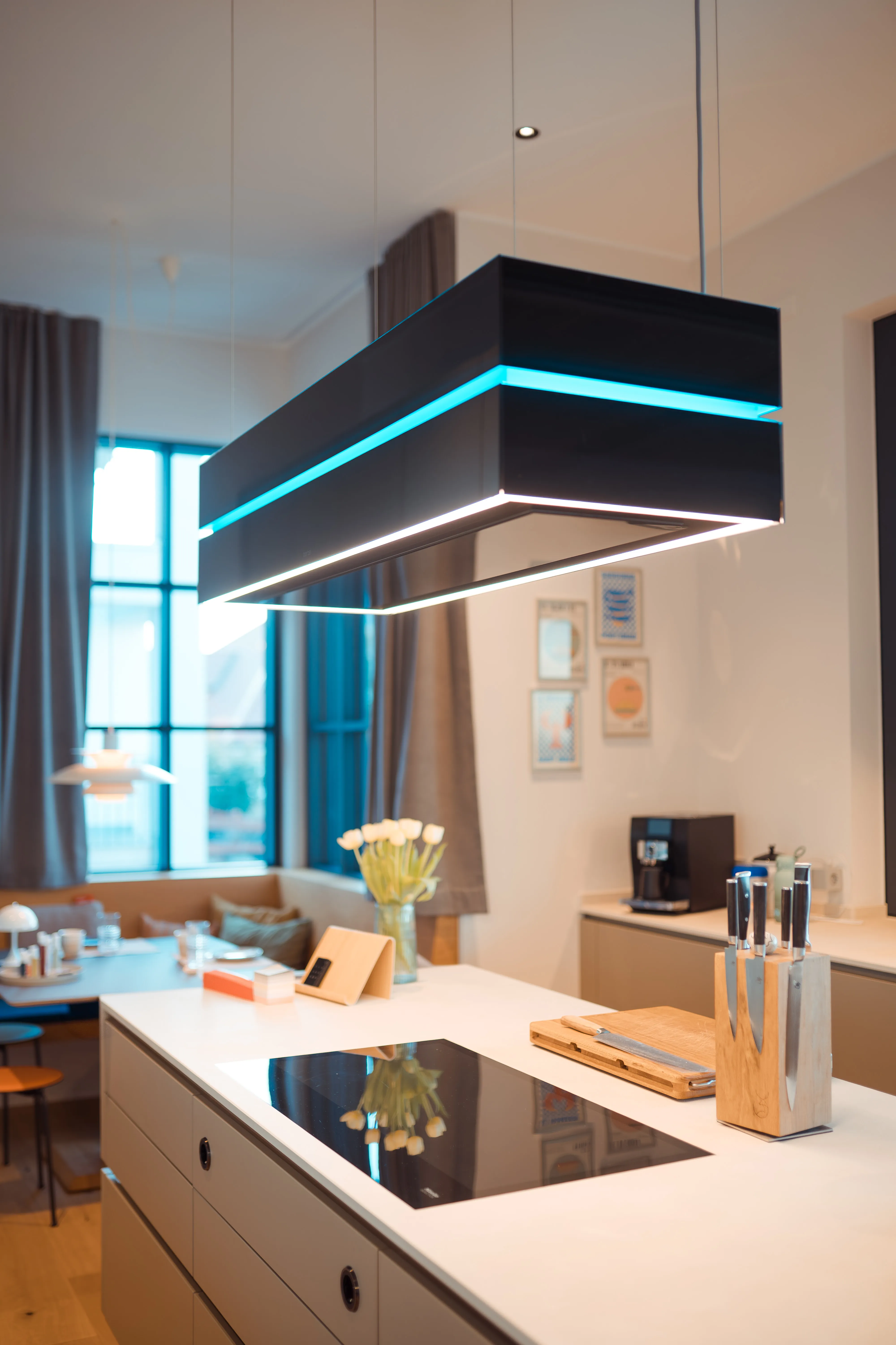 Moderne Küche mit Kochinsel, schwarzer Induktionskochfeld, Messerblock mit Messern, Holzbrett und hängender rechteckiger Deckenlampe mit blau-weißer LED-Beleuchtung, im Hintergrund Esstisch mit Stühlen und Fenster mit Vorhängen.