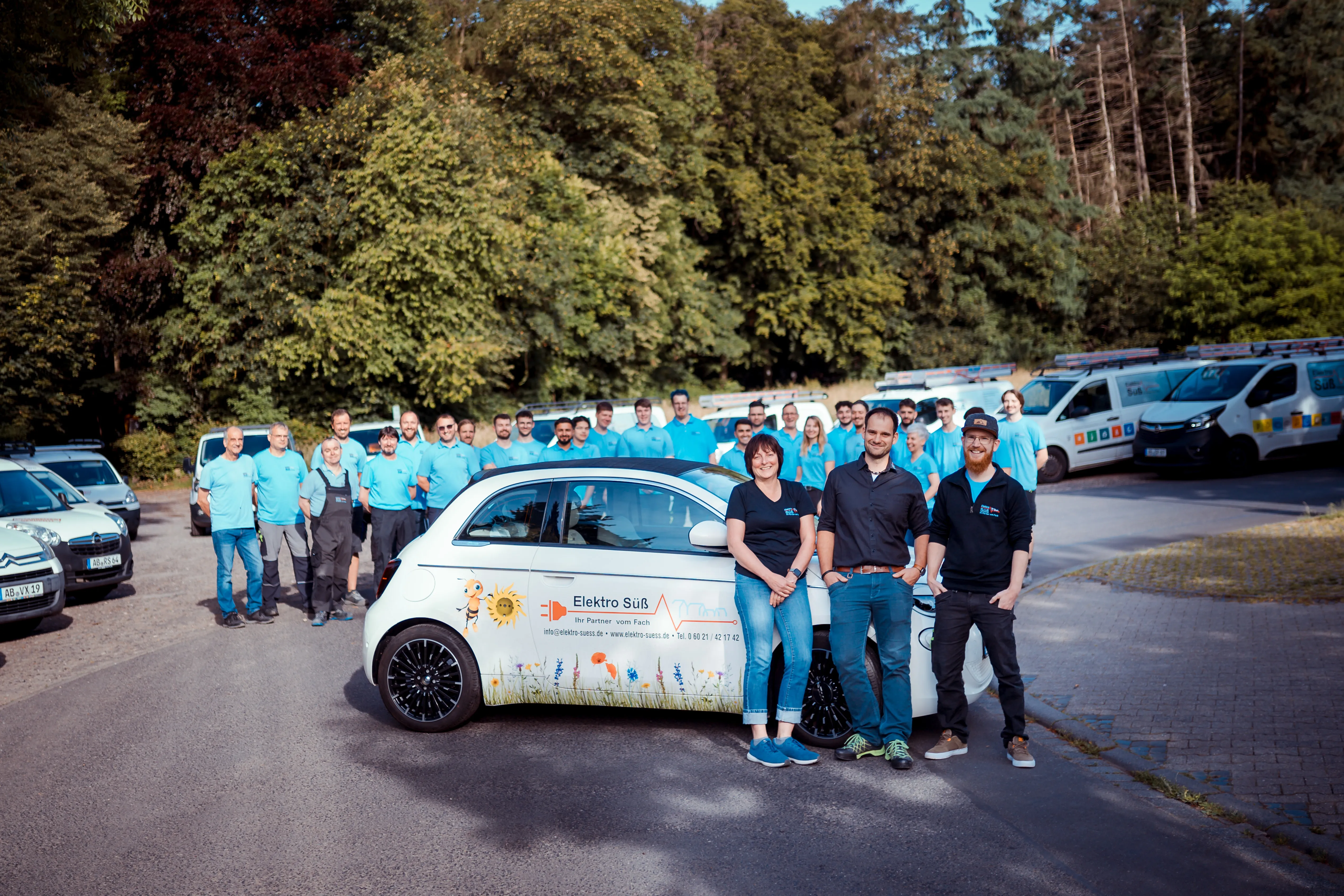 Gruppenfoto neben einem Auto mit Elektro Süß Logo in der Mitte und mehreren Firmenfahrzeugen im Hintergrund, umgeben von Bäumen.