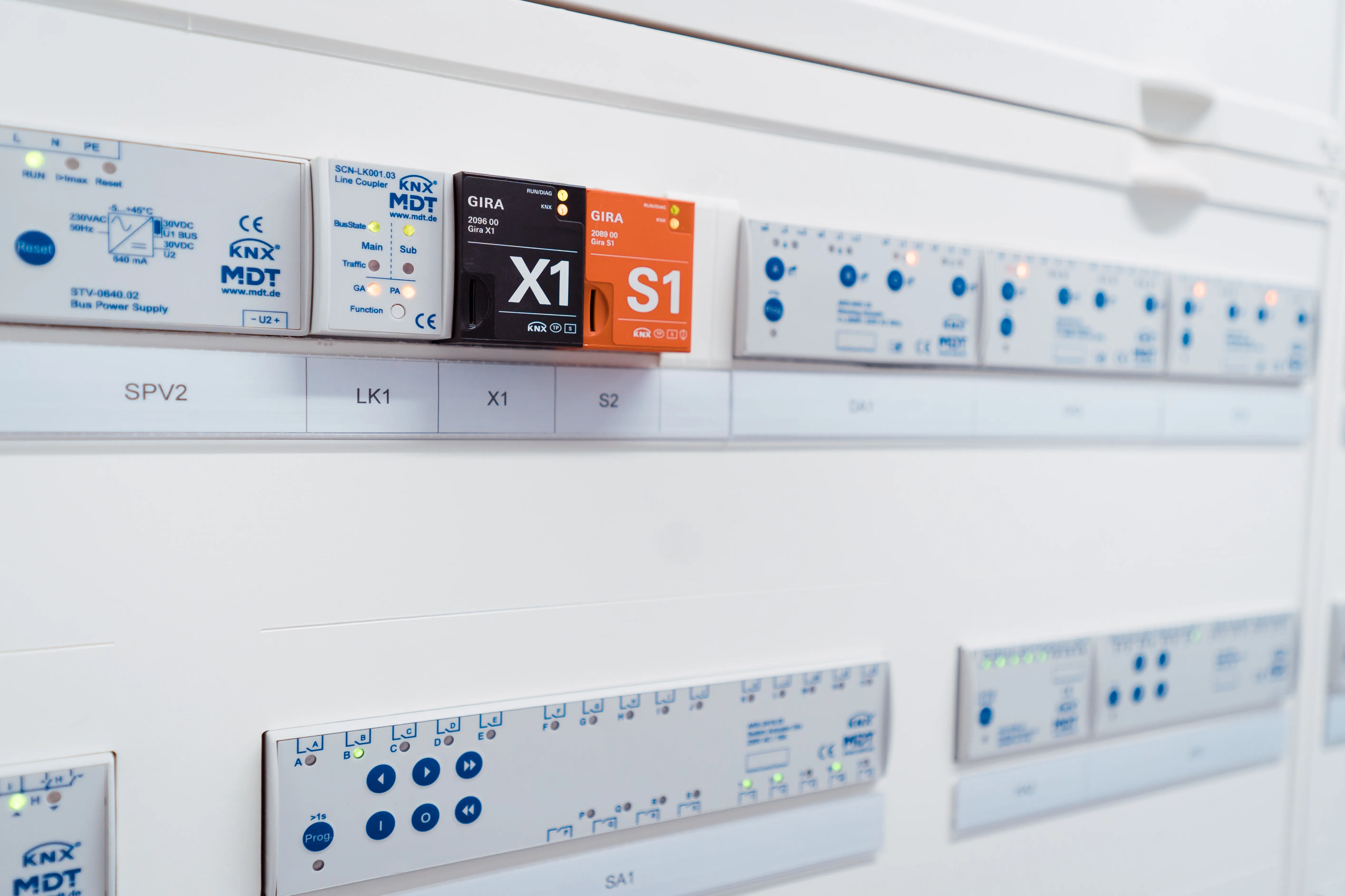 Nahaufnahme einer Elektroanlage mit beschrifteten Modulen und Leuchtanzeigen, darunter GIRA Module mit Markierungen X1 und S1 sowie MDT KNX-Komponenten.