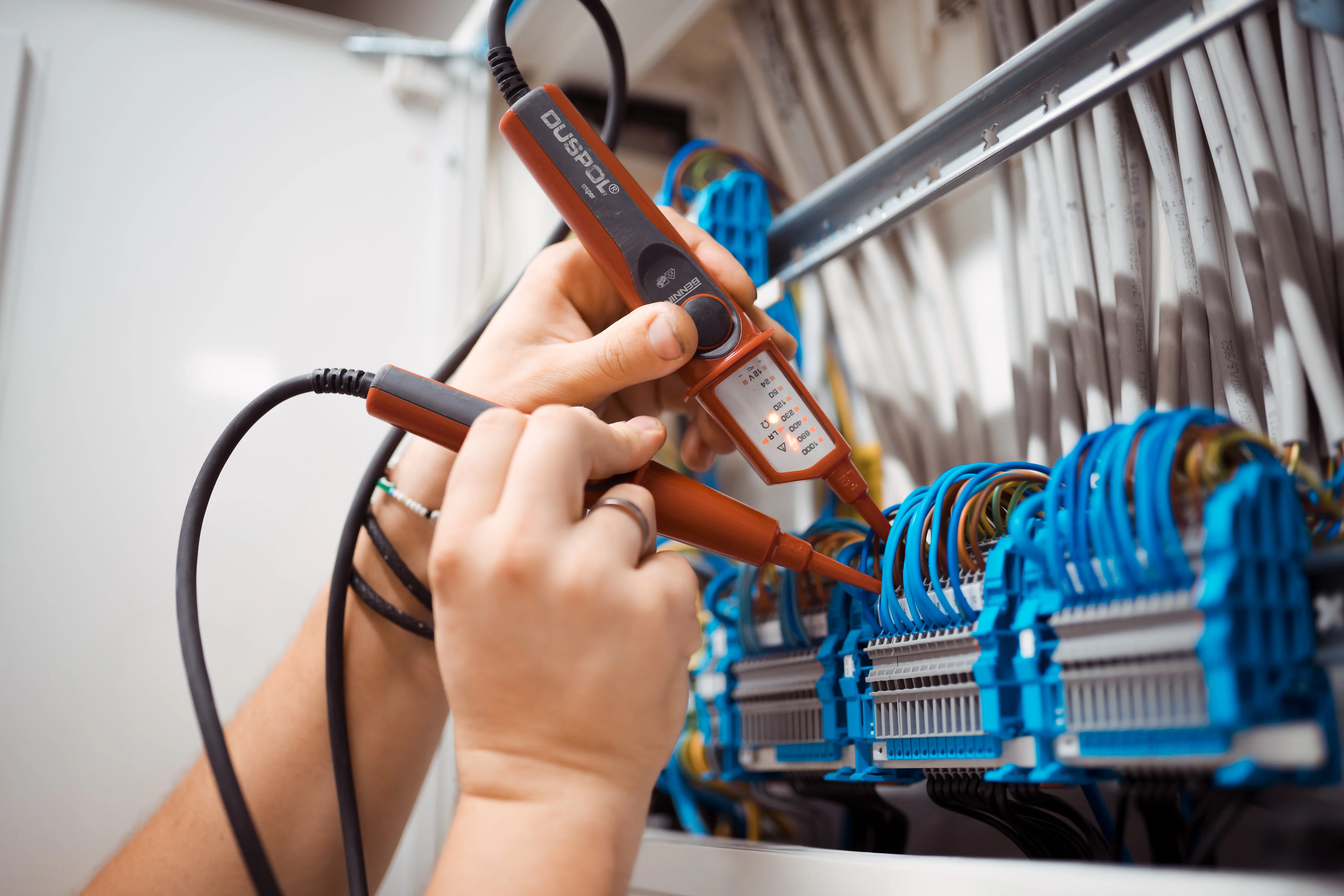 Eine Person prüft elektrische Verbindungen mit einem voltmeterähnlichen Messgerät an einem Schaltschrank mit zahlreichen blauen und grauen Kabeln und Klemmen.