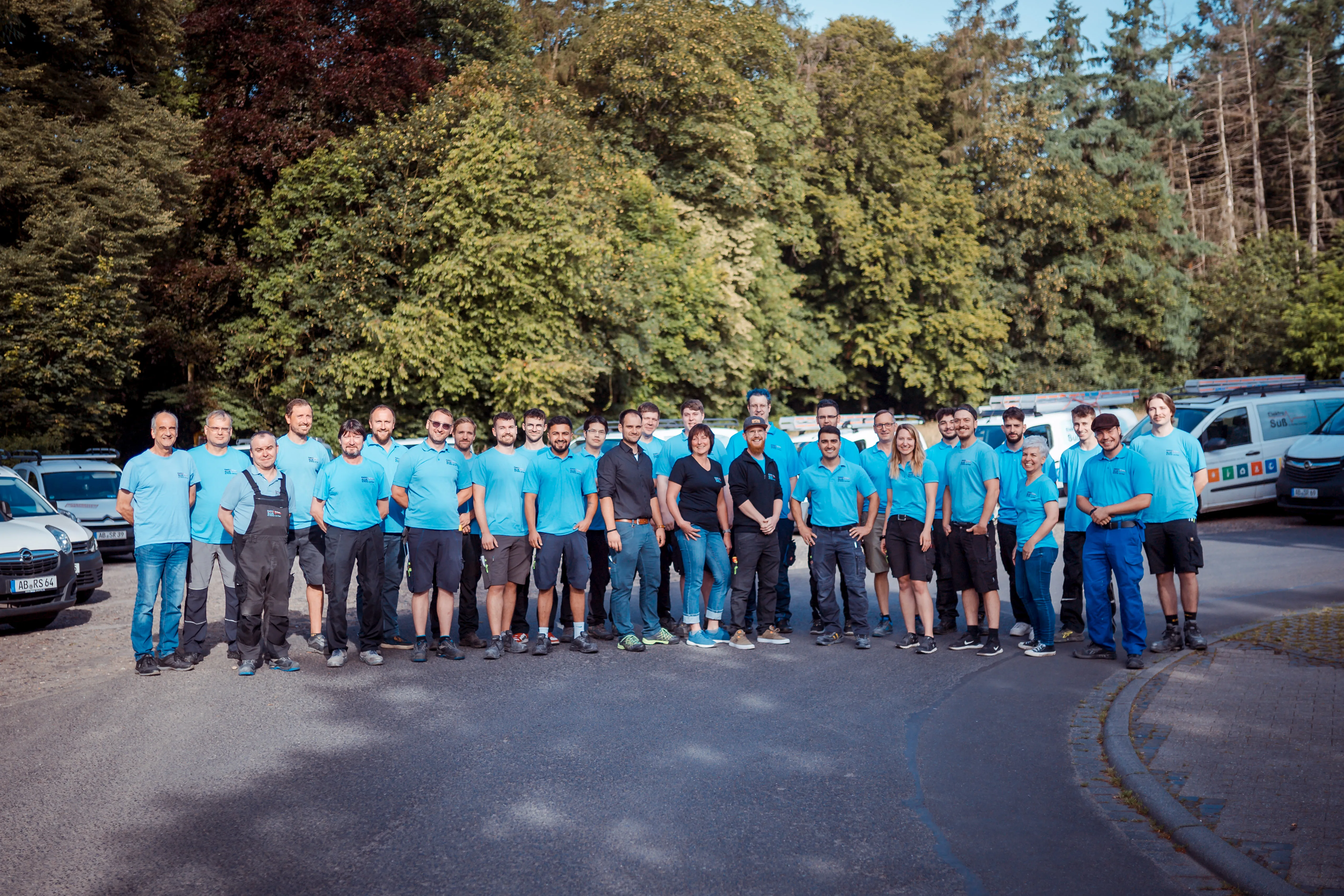 Eine große Gruppe von Erwachsenen, die in hellblauen Firmen-T-Shirts und Arbeitskleidung gekleidet sind, steht auf einer Straße vor großen grünen Bäumen; im Hintergrund sind Firmenfahrzeuge zu sehen.