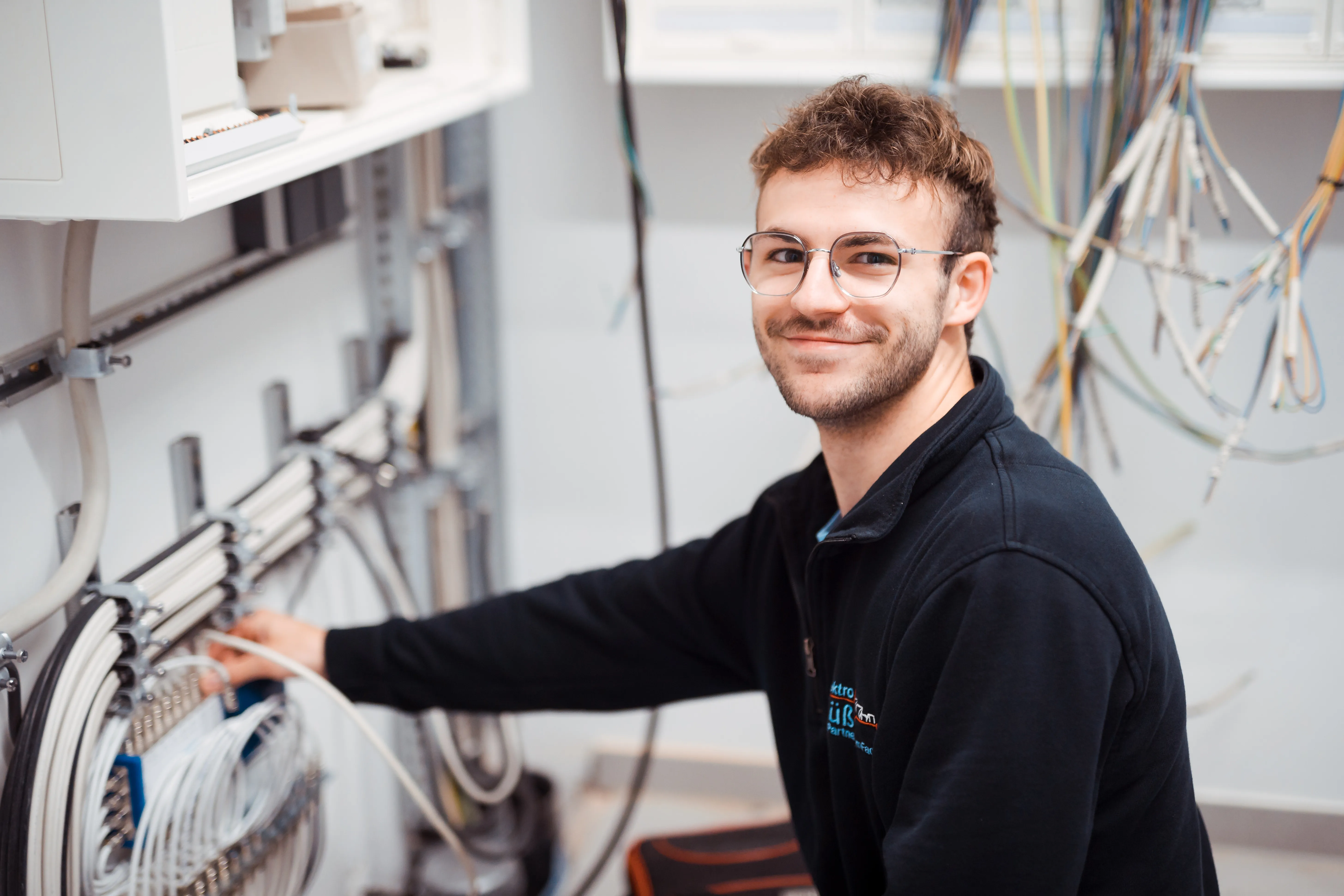 Junger Mann mit Brille und dunklem Pullover arbeitet an einem Schaltkasten und hält ein Kabel in der Hand, er lächelt zur Kamera.