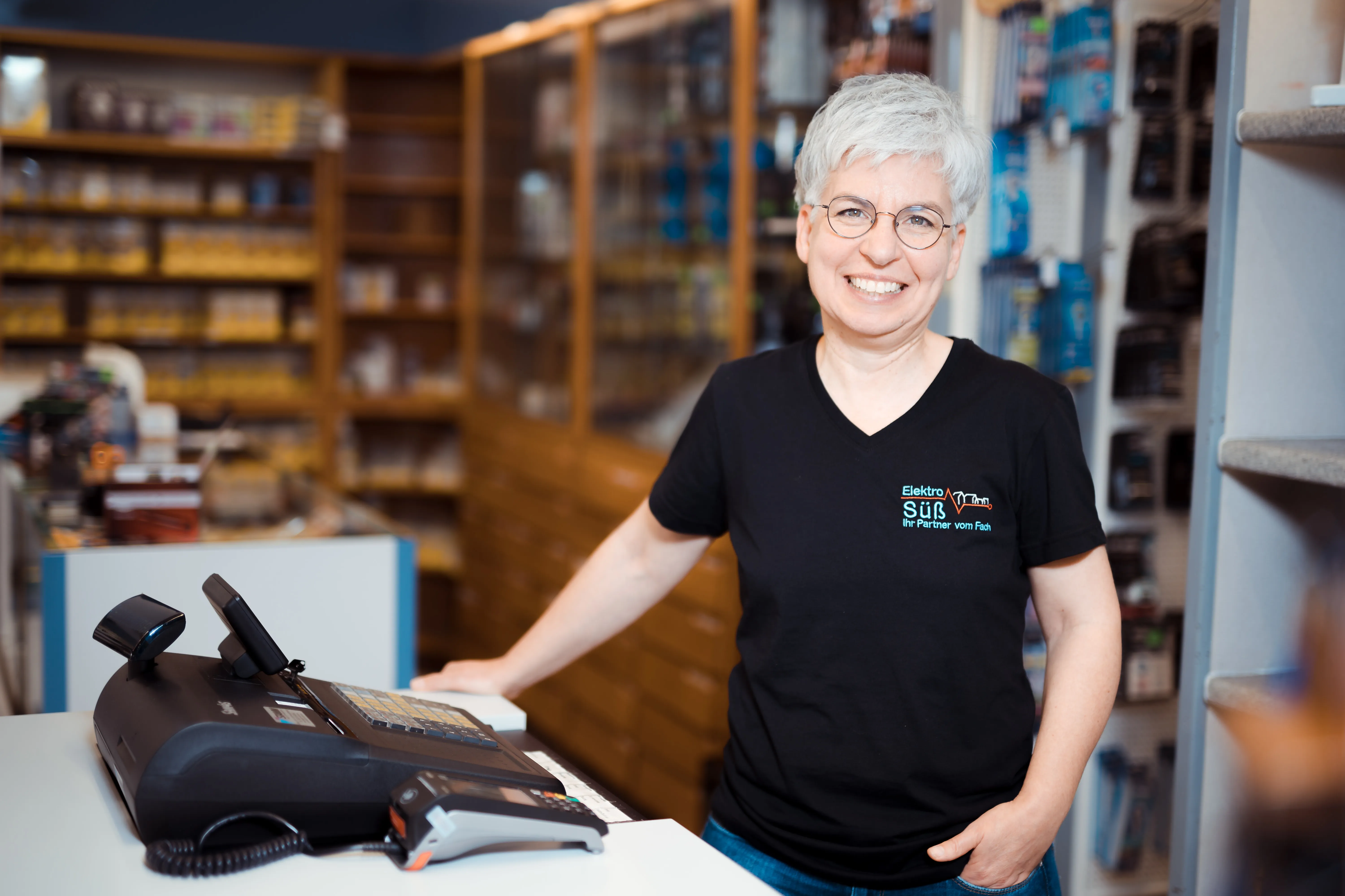 Lächelnde Frau mit kurzen weißen Haaren und Brille steht an einer Ladentheke mit Registrierkasse in einem Geschäft mit Elektroartikeln.