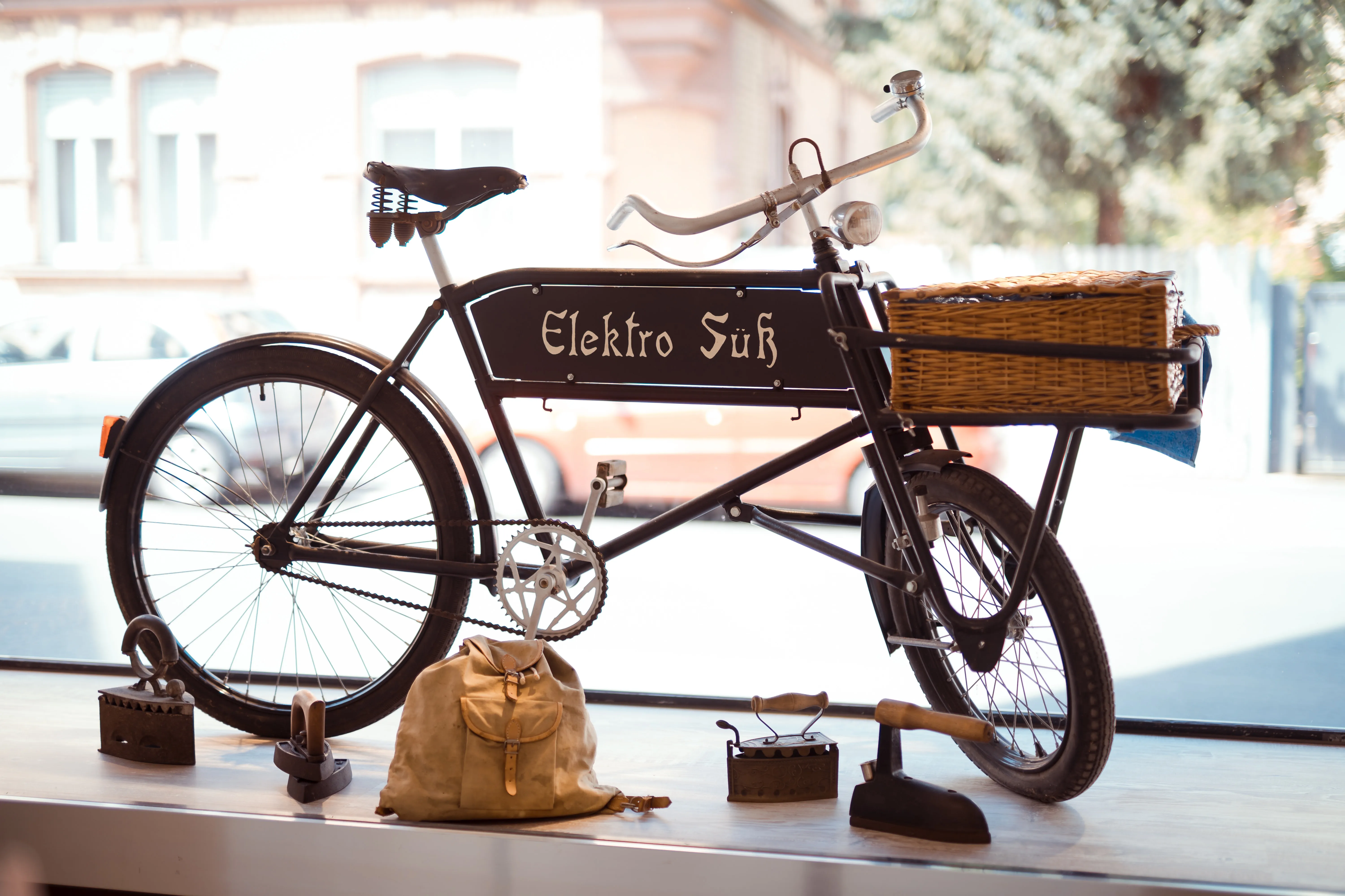 Schwarzes Vintage-Fahrrad mit Korb vorne und Schriftzug Elektro Süß, auf einem Fensterbrett ausgestellt, umgeben von antiken Bügeleisen.