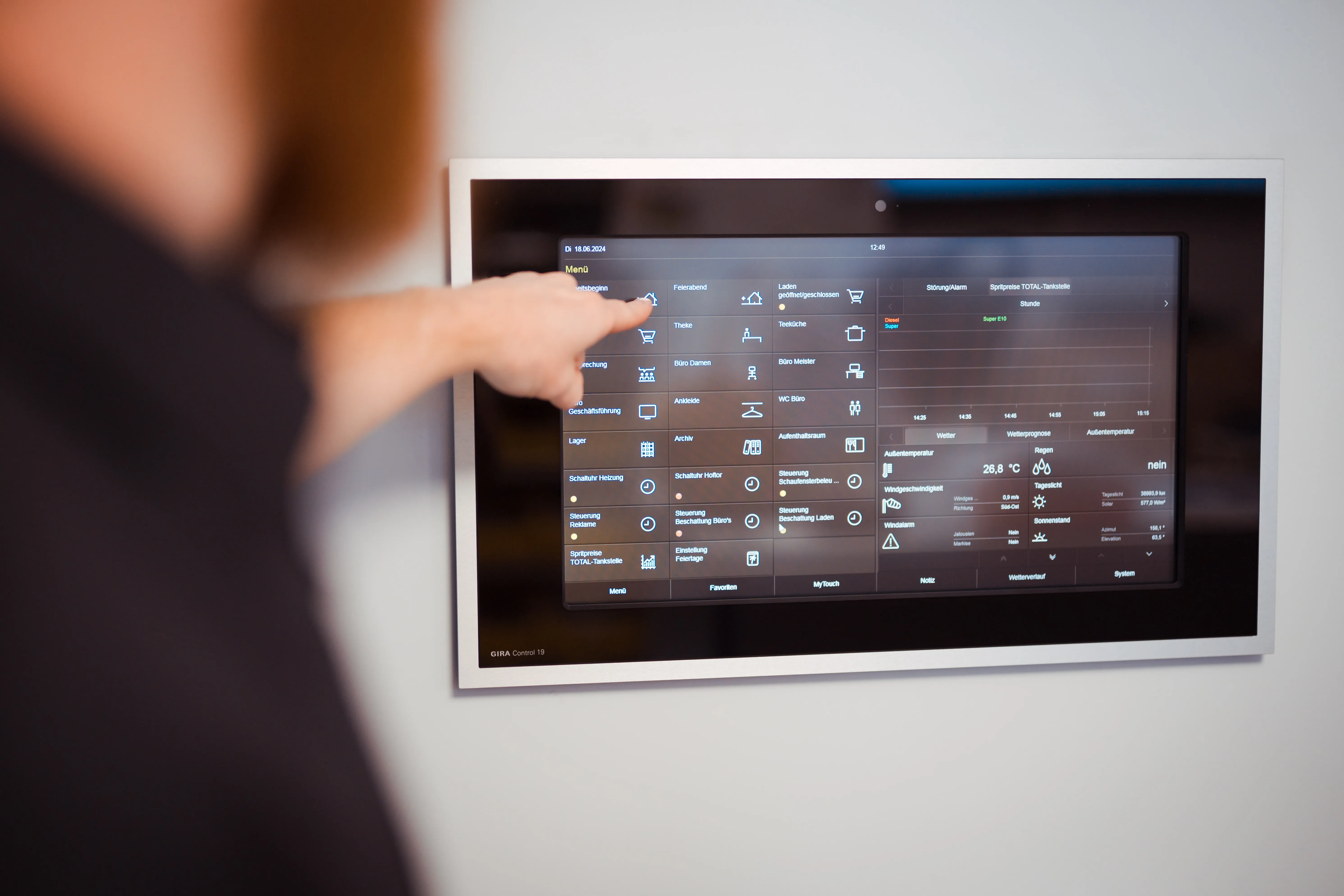 Hand zeigt auf ein Wandsteuerungsgerät mit Touchscreen, das verschiedene Menu-Optionen und Informationen wie Temperatur, Wetter und Steuerungen anzeigt.