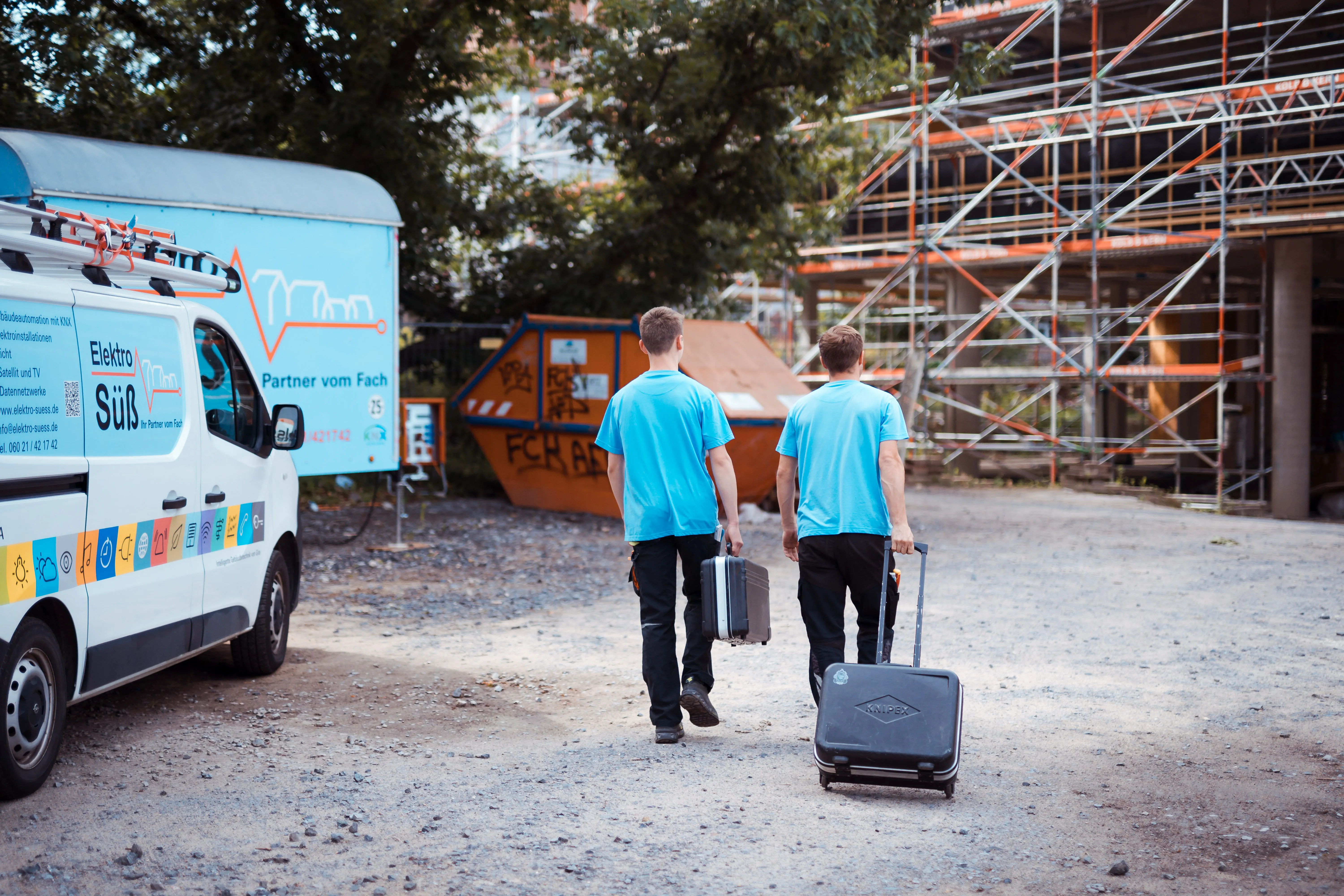 Zwei Männer in hellblauen T-Shirts gehen auf eine Baustelle zu, tragen Werkzeuge in Koffern, links steht ein weißer Van mit der Aufschrift Elektro Süß, im Hintergrund Gerüste, ein blauer Anhänger und ein oranger Container.