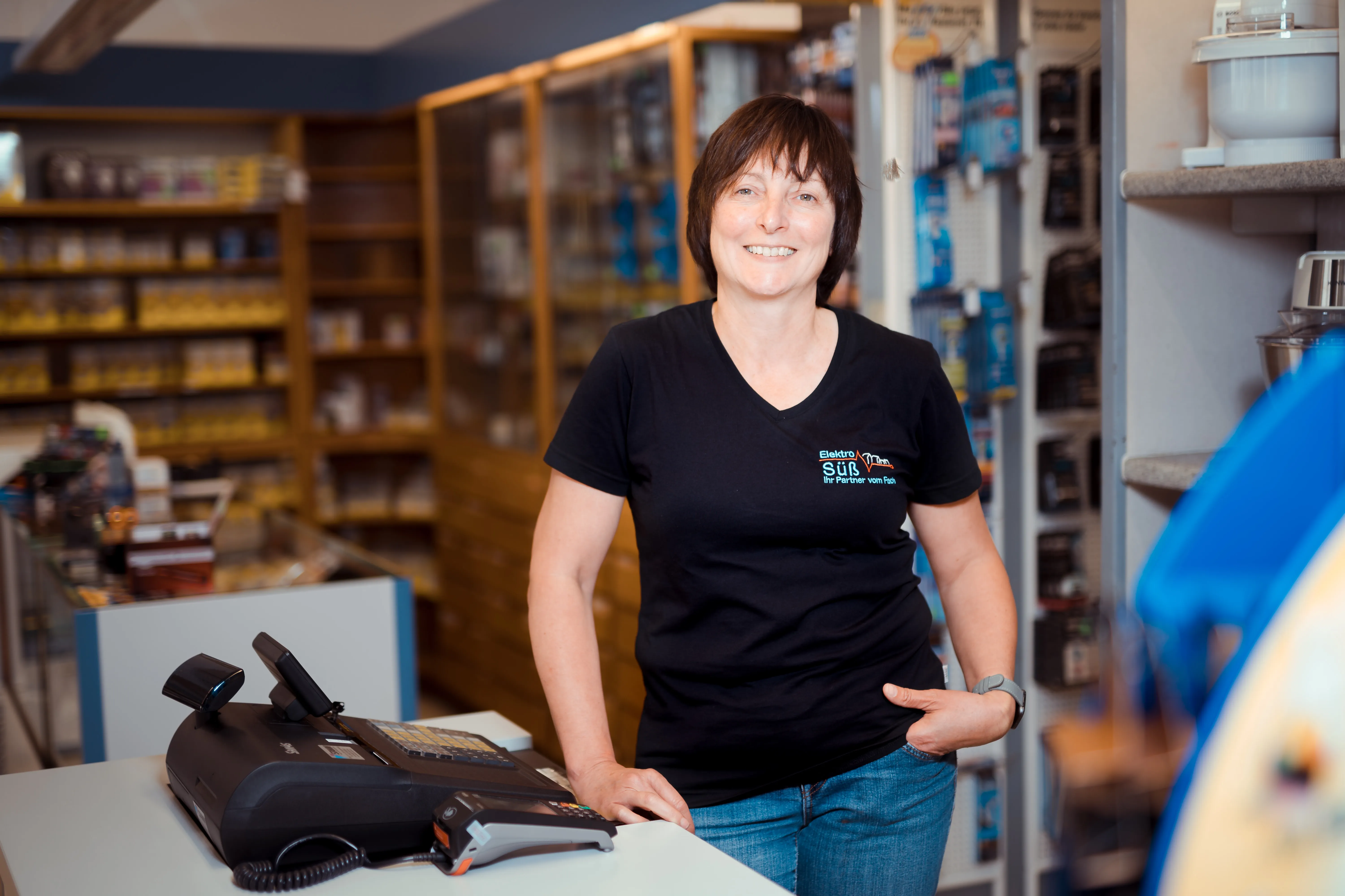 Frau in schwarzem T-Shirt mit Firmenlogo lehnt am Ladentisch neben einer Registrierkasse und lächelt in einem Geschäft mit Regalen voller Produkte im Hintergrund.