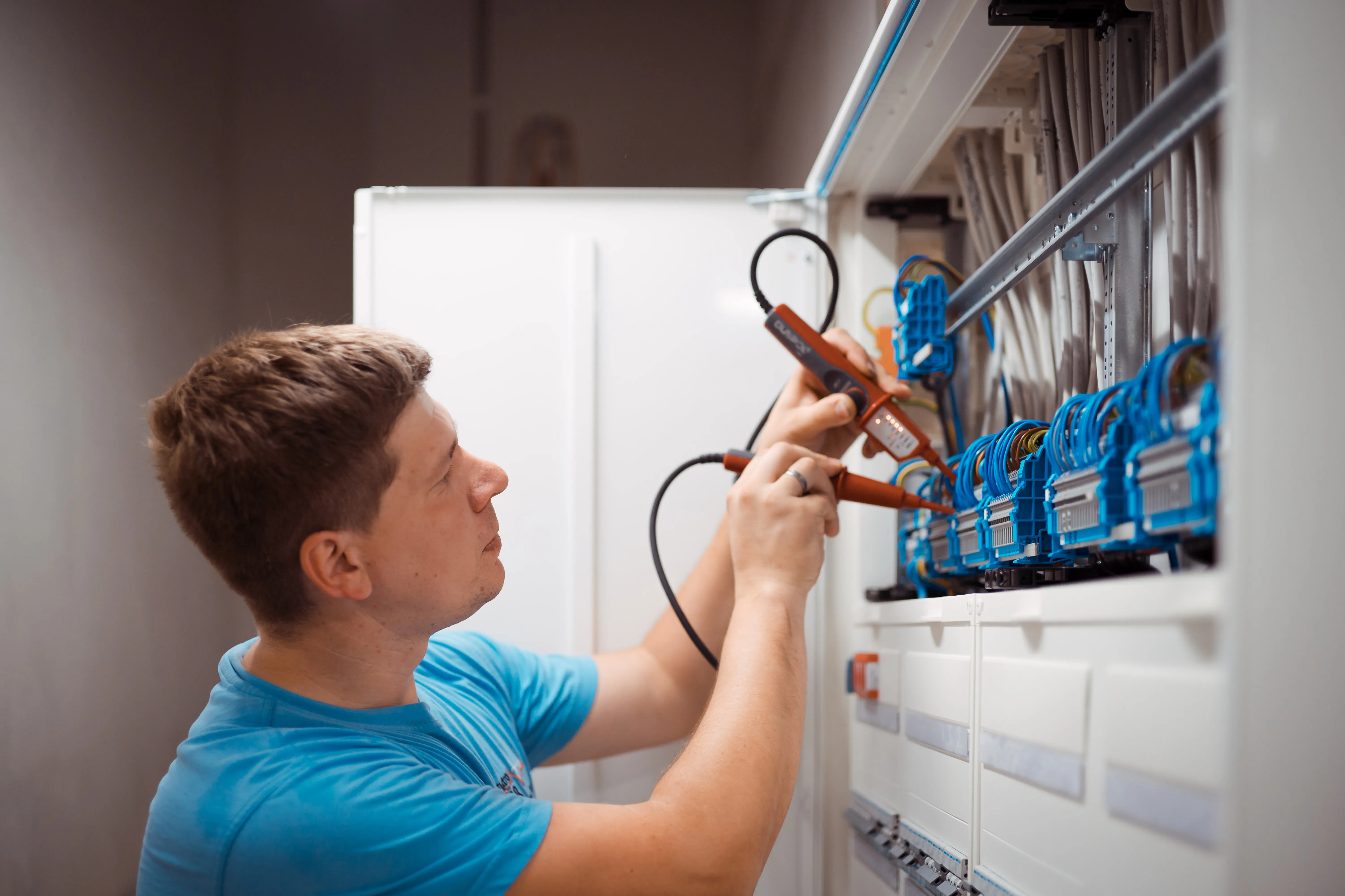 Ein Elektriker in einem hellblauen T-Shirt prüft mit Messgeräten die Verkabelung in einem elektrischen Schaltschrank mit blauen Kabeln und Anschlüssen.