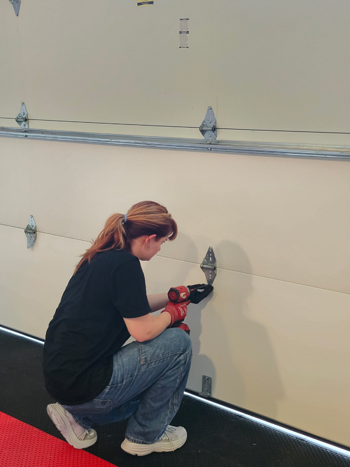 Person with red gloves using a red drill to fix a beige garage door while kneeling on a black floor mat.