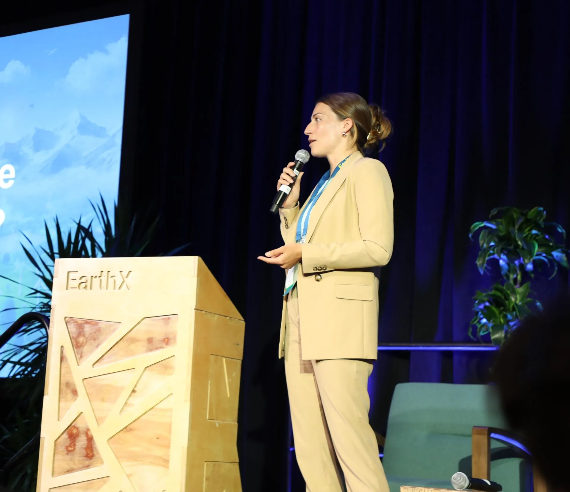 A woman in a beige suit stands on stage holding a microphone, speaking near a wooden podium with the EarthX logo, with plants and a blue curtain in the background.