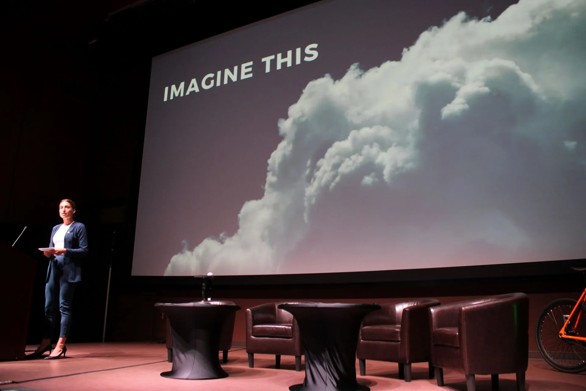 A woman in a blue suit stands at a podium on stage next to round tables and brown leather chairs, with a large screen behind her displaying the words 'IMAGINE THIS' and an image of thick white smoke or clouds on a dark background.