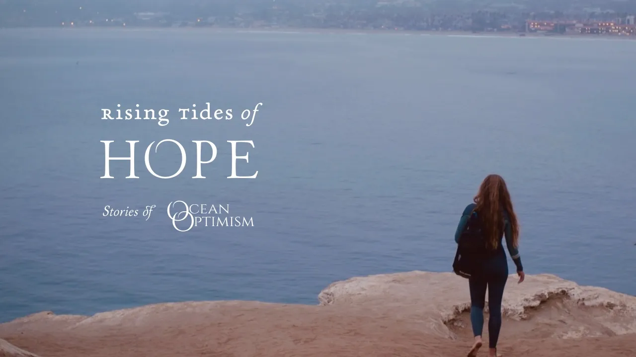 A person with long hair and a backpack walks barefoot on rocky cliffs overlooking a calm ocean with a distant coastline under a cloudy sky, with the text 'Rising tides of HOPE Stories of Ocean Optimism' on the left.