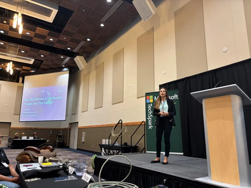 A woman stands on a stage in a conference room beside a podium and a Microsoft TechSpark banner, giving a presentation titled 'Can We Harness AI To Benefit People And The Planet?' displayed on a large screen. Audience members are seated at tables in the foreground.