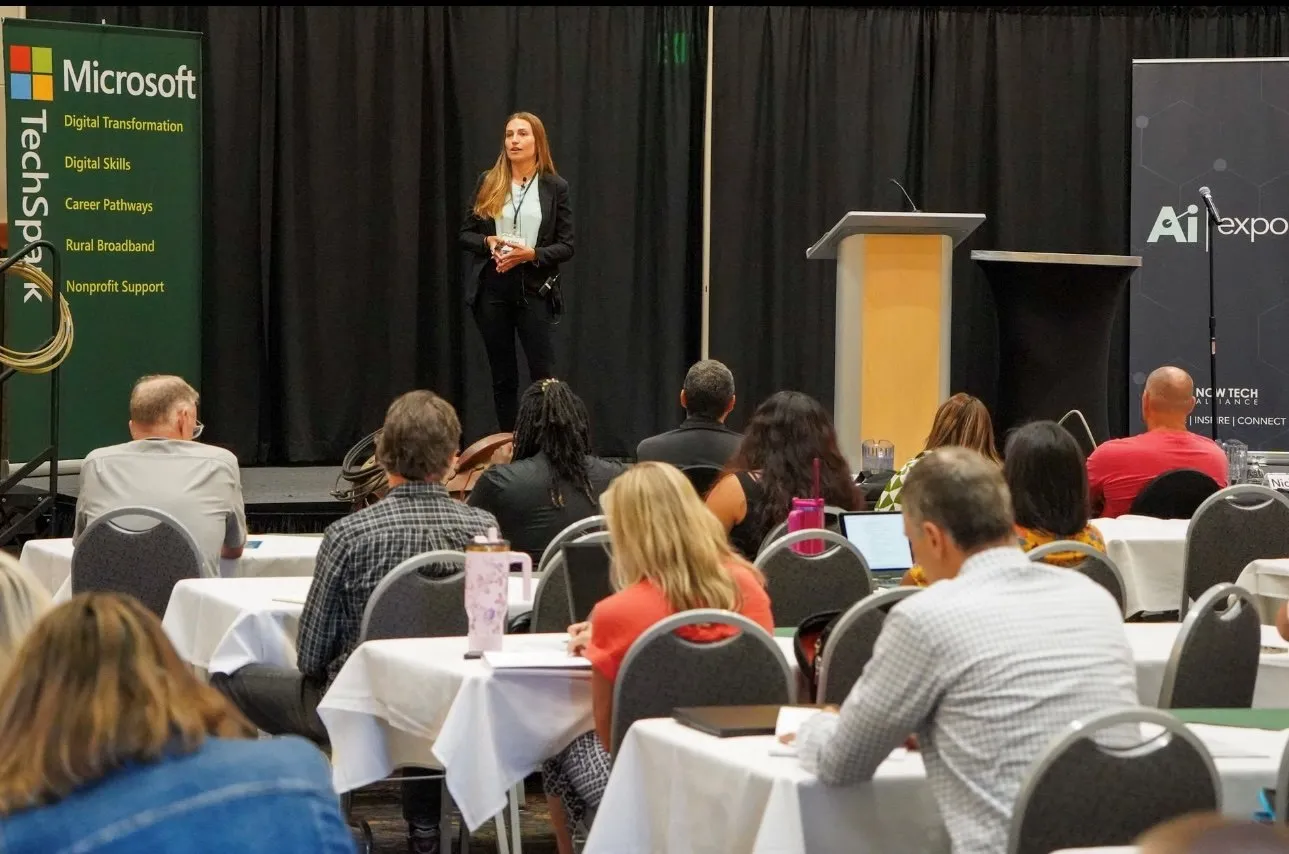 A woman in business attire stands on a stage speaking to an audience sitting at tables with white tablecloths, with Microsoft and AI Expo banners visible behind her.