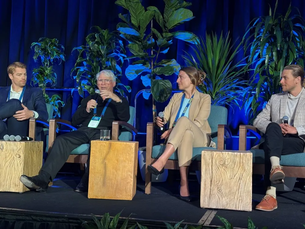 Four panelists seated on stage in blue chairs holding microphones, engaged in a discussion, with large green plants and blue curtains in the background.