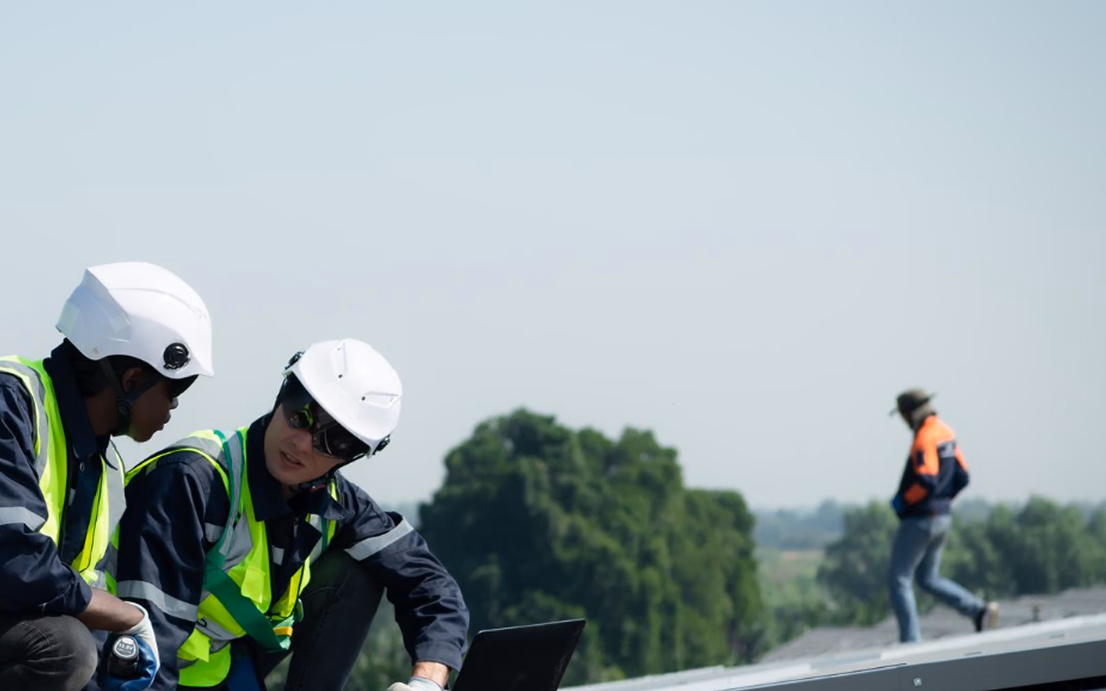 Two construction workers in safety gear looking at a laptop on a rooftop, another worker walks in background.