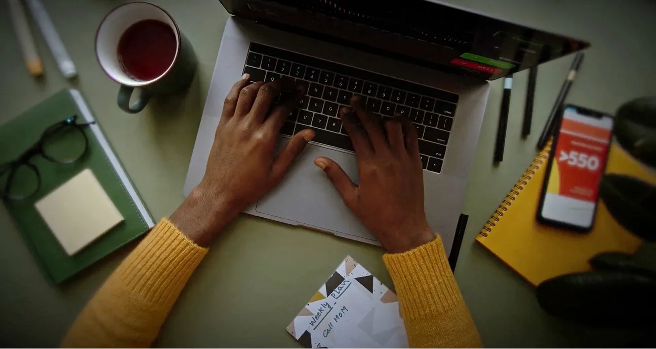 A person typing on a laptop with a cup of tea, notebook, and smartphone nearby on a desk.