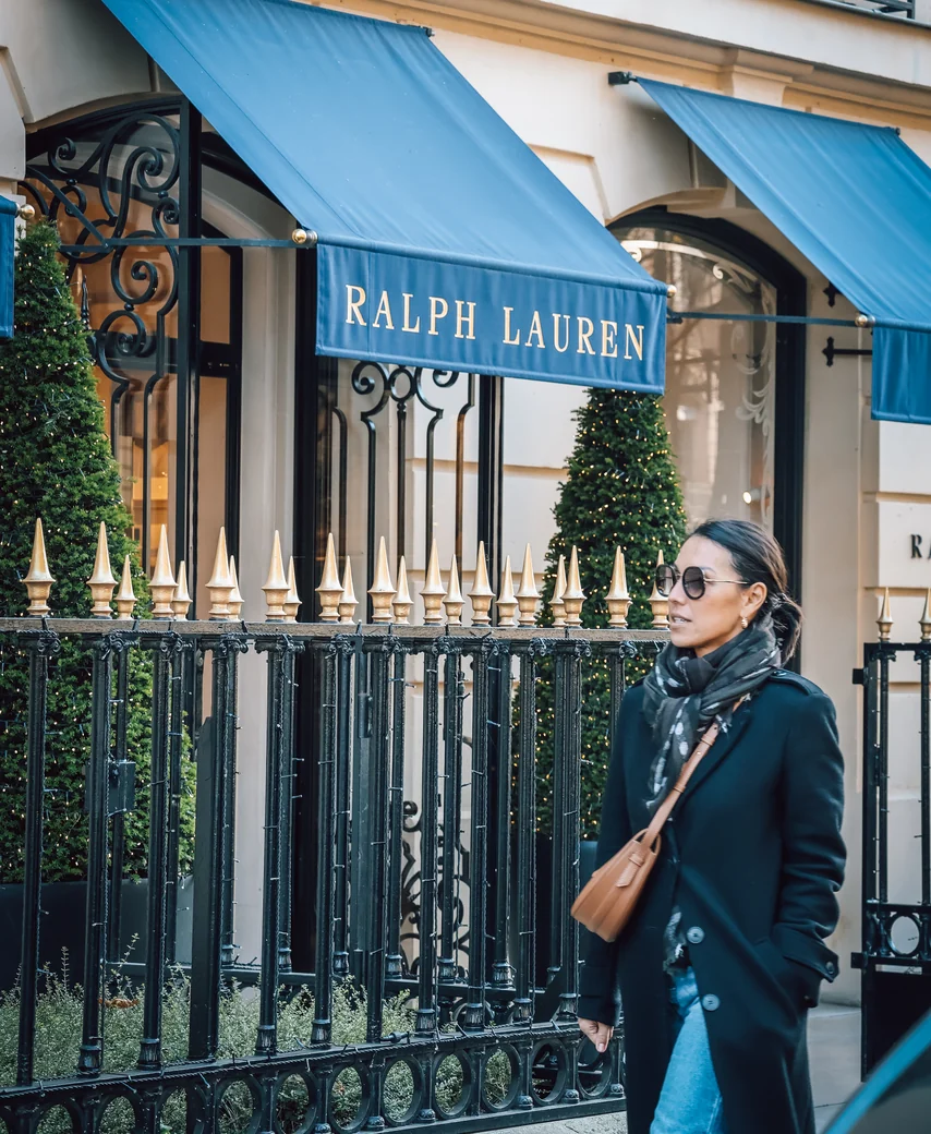 Une femme portant un manteau noir, une écharpe et des lunettes de soleil marche devant une boutique Ralph Lauren avec des stores bleus et une grille noire ornementale.