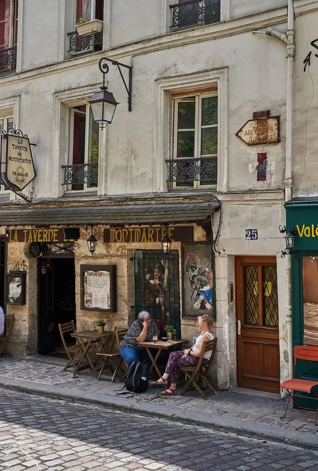 Deux femmes sont assises à une table en bois à l'extérieur de la taverne La Taverne de Montmartre, située dans un bâtiment ancien avec des fenêtres ornées de balustrades en fer forgé et une enseigne suspendue, au numéro 25 d'une rue pavée.