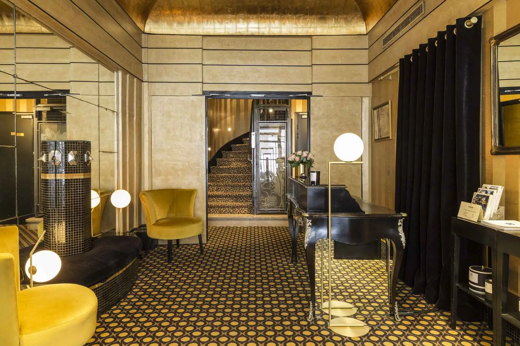 Réception élégante d'hôtel avec tapis à motifs circulaires jaunes et noirs, deux fauteuils jaunes, et un bureau noir vintage avec une lampe sphérique. Un escalier en colimaçon avec tapis assorti est visible à l'arrière-plan. Les murs sont décorés de miroirs et rideaux noirs.