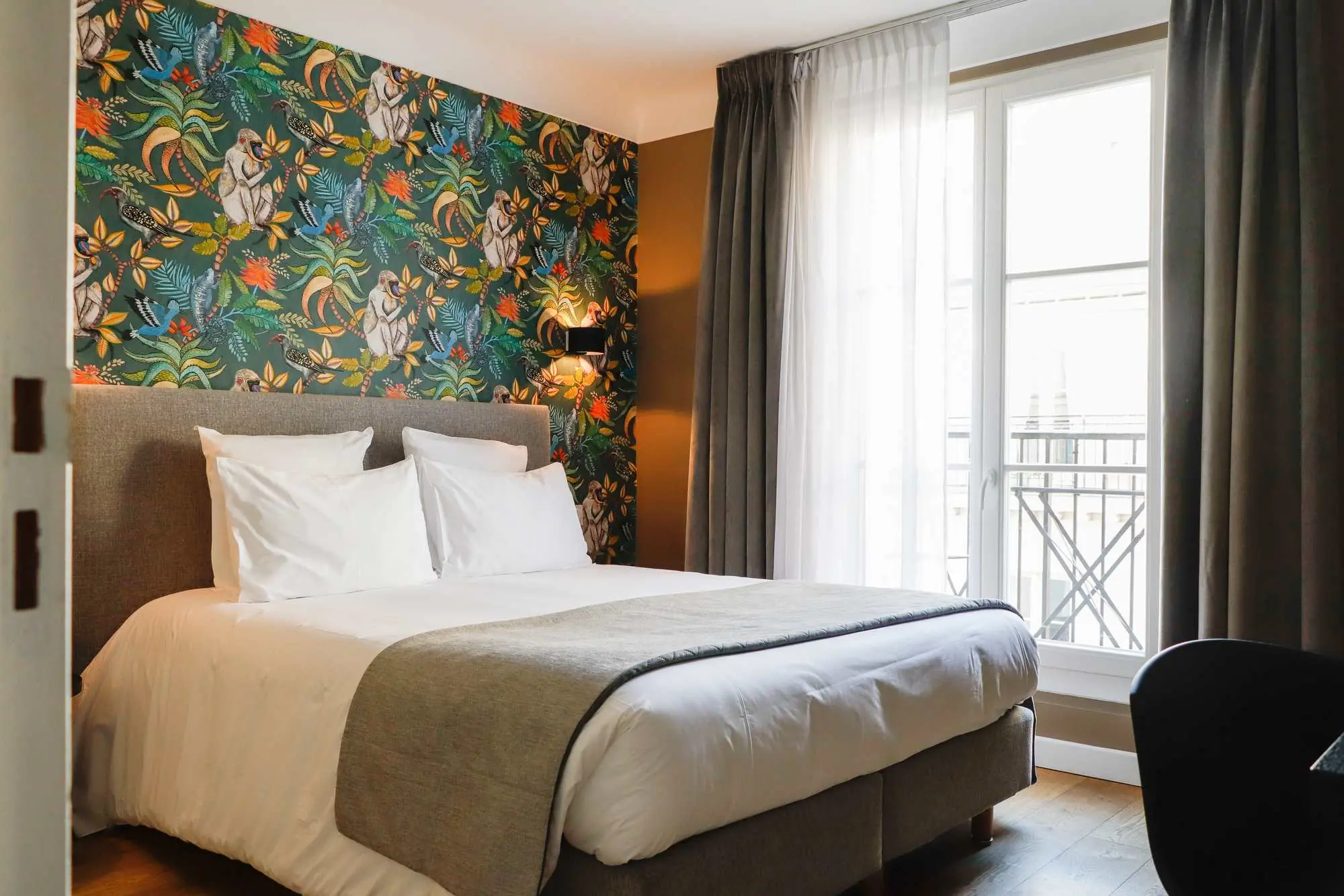 Chambre d'hôtel avec un grand lit double blanc décoré de quatre oreillers, une couverture grise au bout et un mur d'accent avec un papier peint coloré illustrant des singes et de la végétation tropicale, ainsi que de grandes fenêtres avec rideaux gris et transparents laissant entrer la lumière naturelle.