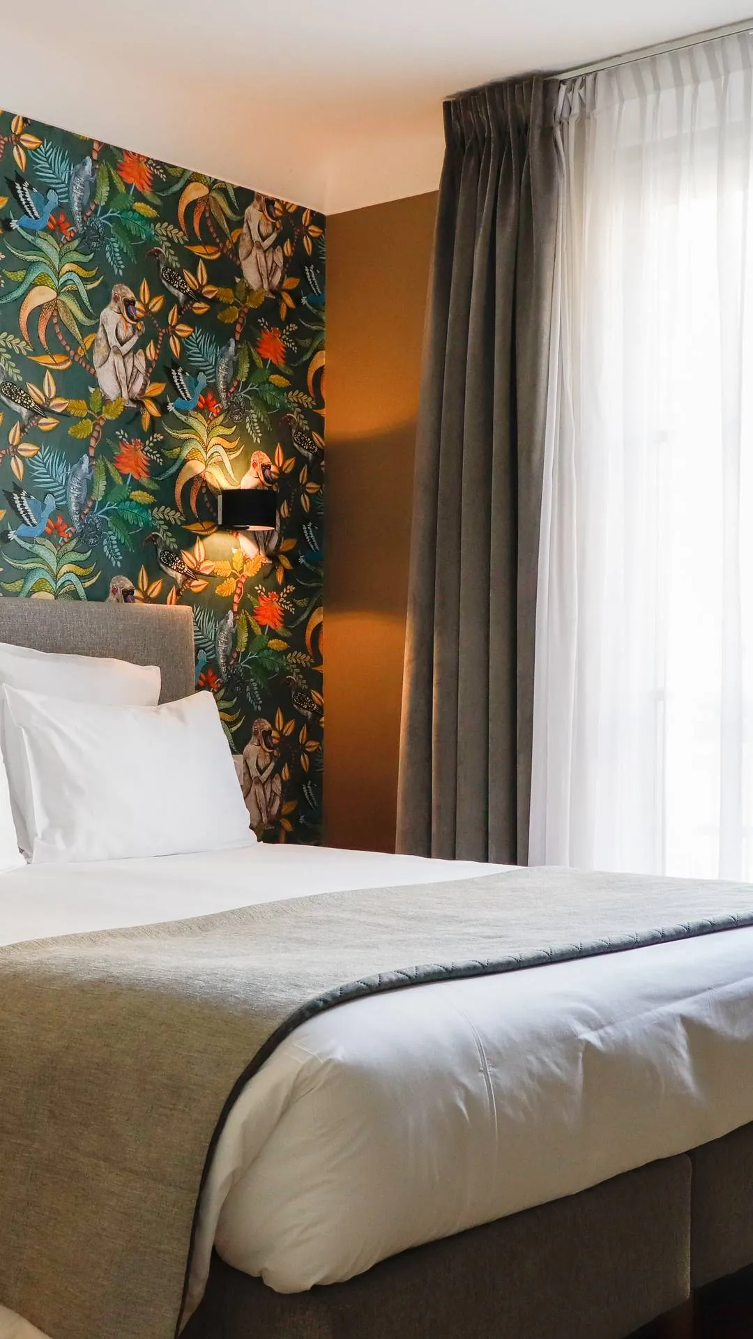 Chambre d'hôtel avec un lit double bien fait, draps blancs, couverture beige, tête de lit grise, mur décoré avec un papier peint coloré à motifs d'oiseaux et de singes, applique murale allumée, et rideaux épais gris à côté d'un rideau translucide blanc.