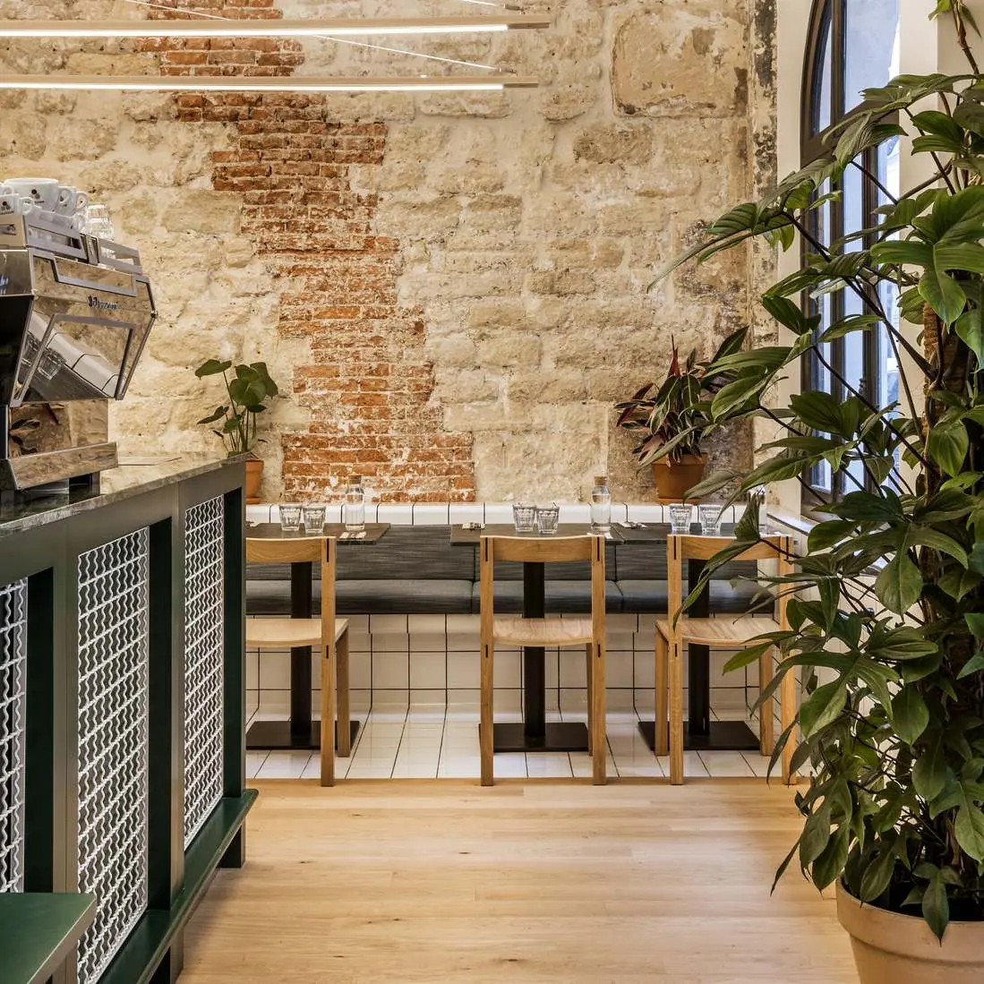 Intérieur d'un café au style moderne et rustique avec murs en pierre apparente, tables et chaises en bois clair, banquettes grises, grandes fenêtres, plantes vertes en pots, et une machine à café argentée sur un comptoir vert avec carreaux blancs.