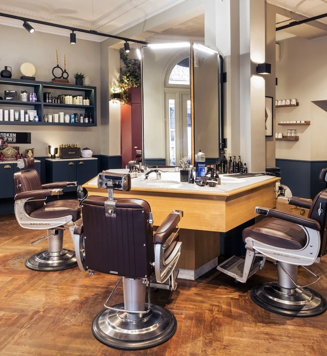 Salon de barbier avec trois fauteuils en cuir marron autour d'un grand miroir éclaire, un comptoir en bois avec produits de soin et outils de coiffure au centre, étagères murales avec produits et décorations dans un intérieur lumineux au sol en parquet.