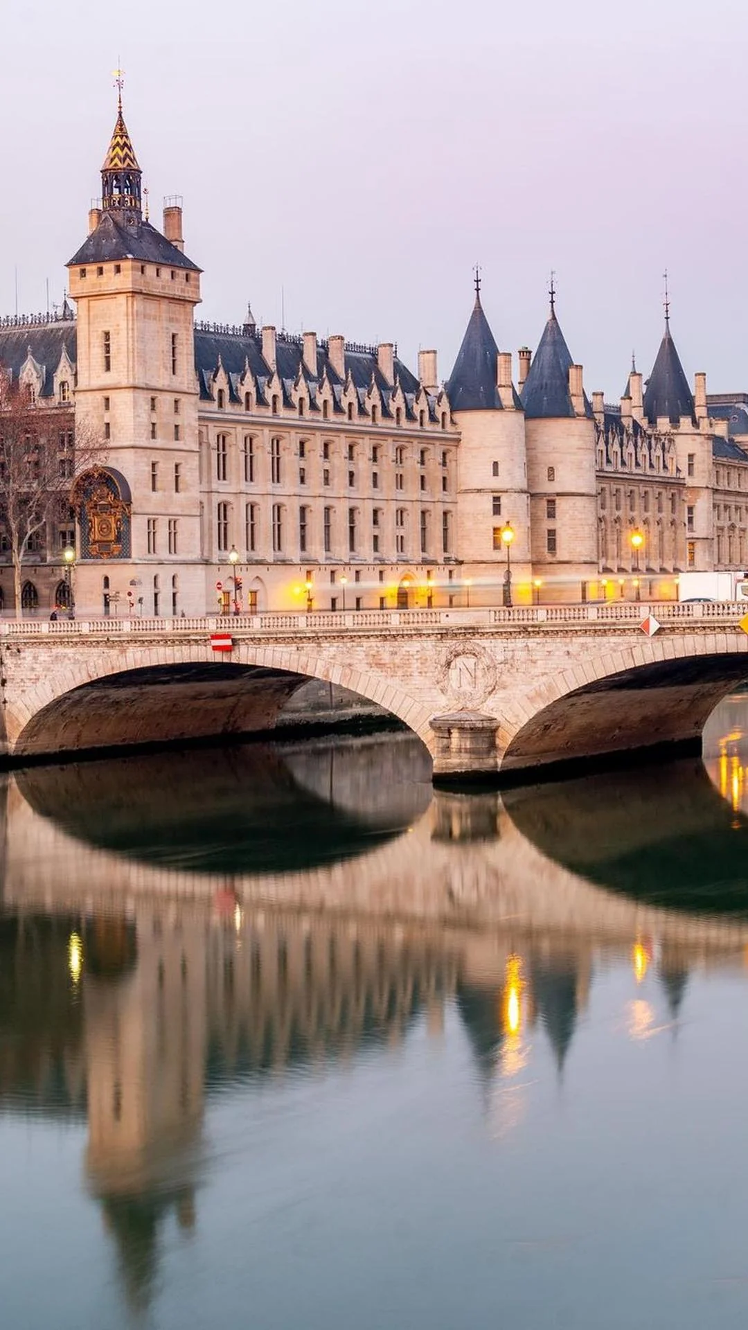 Vue du bâtiment historique de la Conciergerie à Paris avec ses tours en pierre, son toit en ardoise, et un pont en arc qui enjambe la rivière Seine, reflété dans l'eau calme au crépuscule.