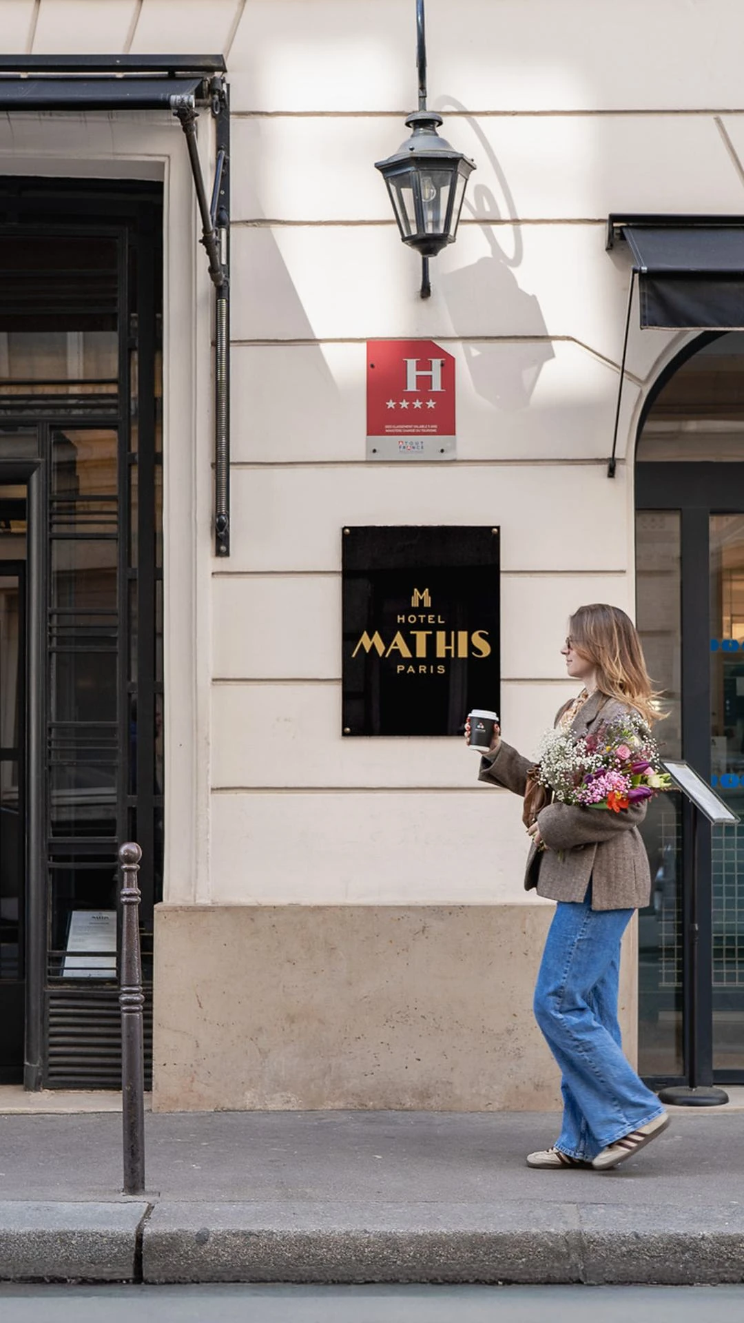 Une femme portant une veste marron, un jean bleu et des baskets tient un bouquet de fleurs colorées et un gobelet de café en marchant devant l'entrée de l'hôtel Mathis à Paris dont le panneau noir avec lettres dorées est visible sur le mur clair, accompagné d'une plaque rouge indiquant une classification hôtelière quatre étoiles.