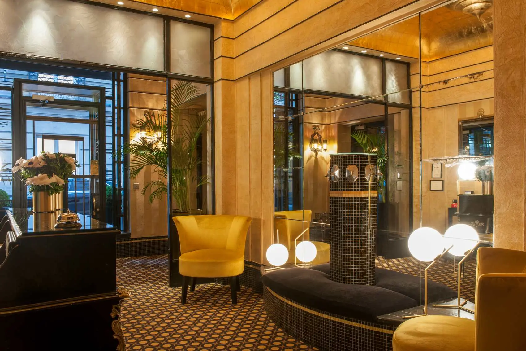 Hall d'entrée d'hôtel luxueux avec des murs dorés, un grand miroir, une banquette noire avec un pied central décoré de carreaux noirs et dorés, deux fauteuils jaunes et des lampes sphériques éclairées.