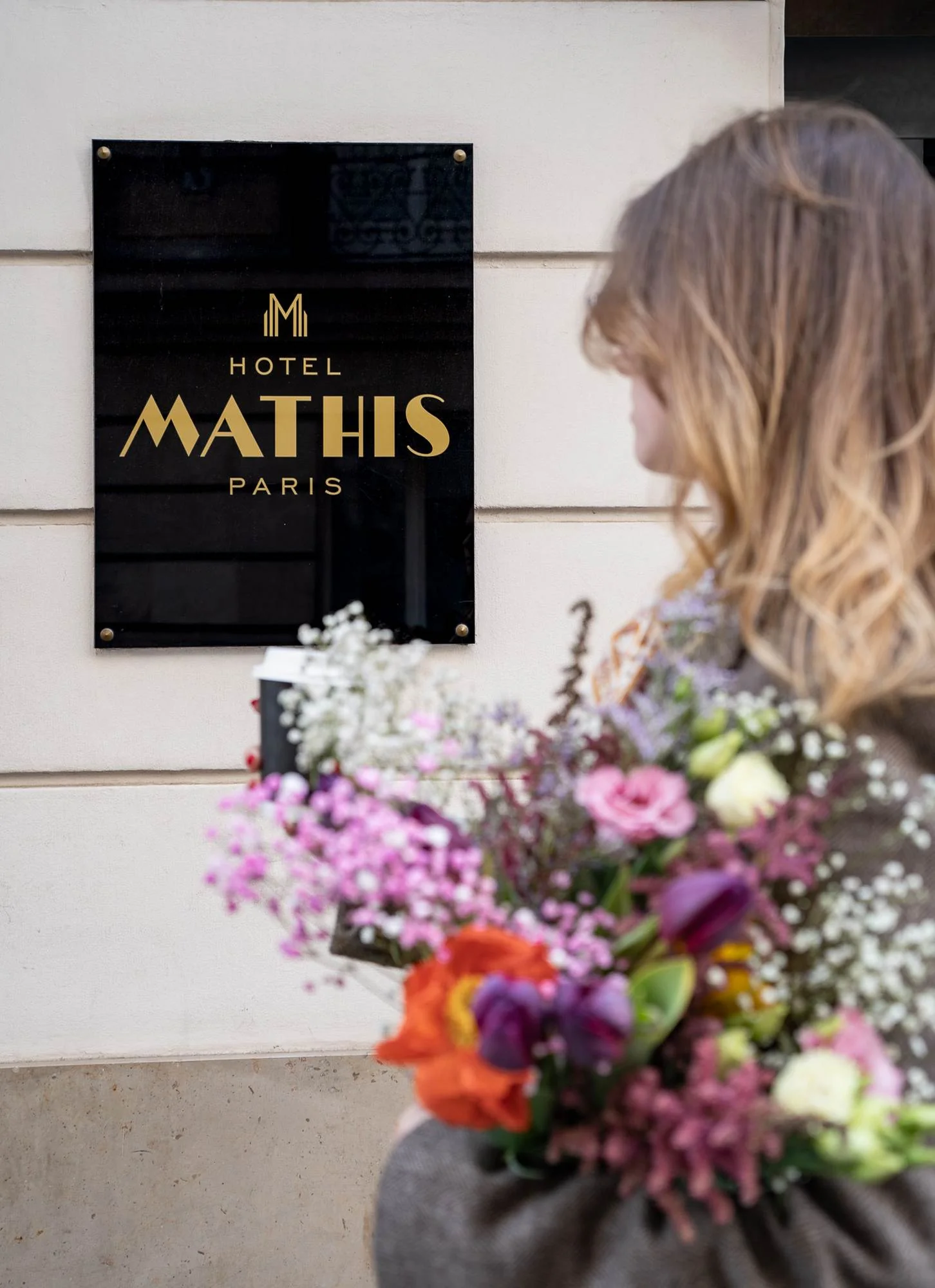 Une femme vue de dos, tenant un bouquet de fleurs colorées, regarde une plaque noire fixée sur un mur beige avec l'inscription dorée « Hotel Mathis Paris ».